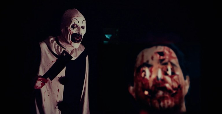 Art the Clown holds a meat cleaver behind a human-head jack-o-lantern in Terrifier (2016).