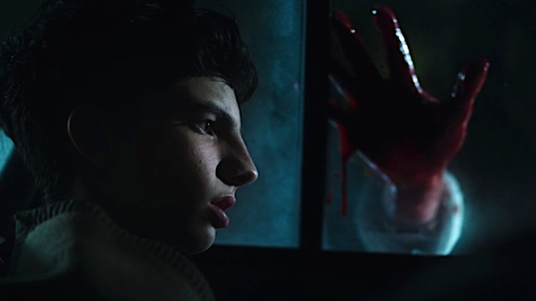 Jair sitting in a car while his possessed mother places a bloody hand on the window in When Evil Lurks (2023).