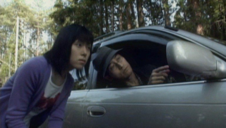 Junji Ito sitting in a car, pointing towards the right as a young woman looks one.