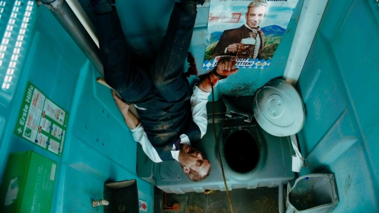 A man scream while upside down in a portable toilet with a metal bar through his left arm in Holy Shit (2022).
