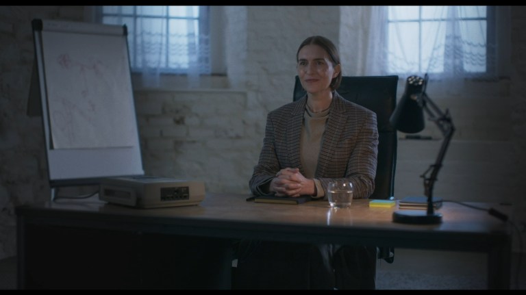 Agnes smiles while sitting at a desk in All you Need is Death (2023).