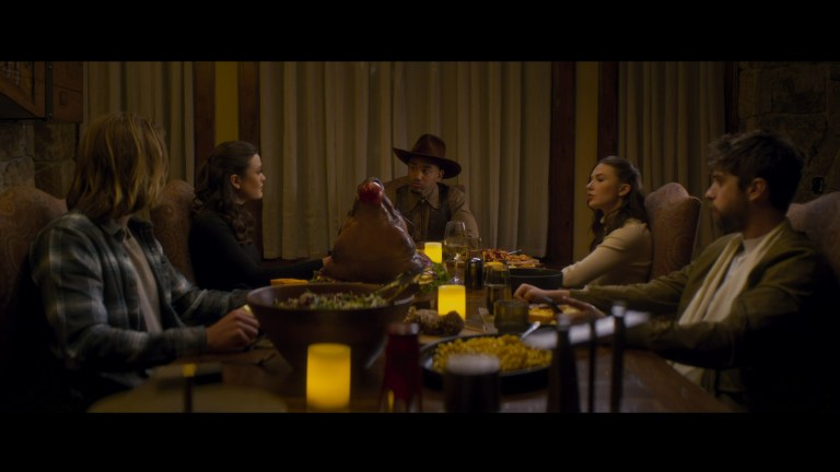 The group gathers at a dinner table with a huge pigs head as the centerpiece in Snow Valley (2024).