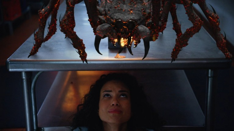 A lava-dripping spider stands on a table as a woman hides underneath in 2 Lava 2 Lantula (2016).