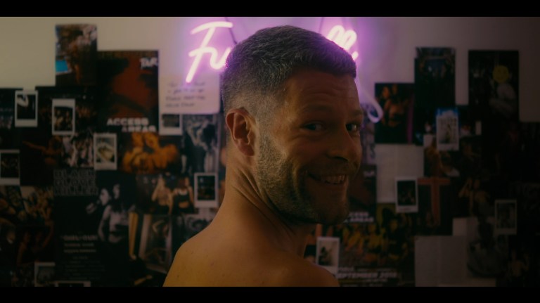 Axel smiles in front of a wall filled with Dakota's photographs in Kill You Lover (2023).