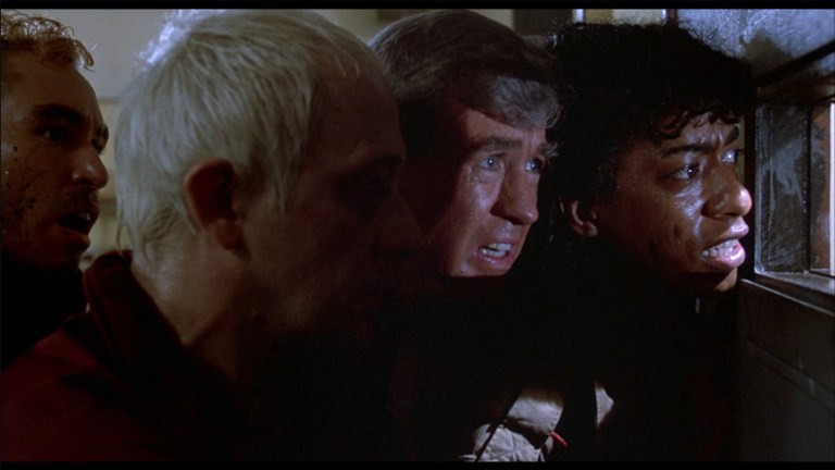 Scuz, Ernie, Burt, and spider peer out of the window in the door of the embalming room in The Return of the Living Dead (1985).