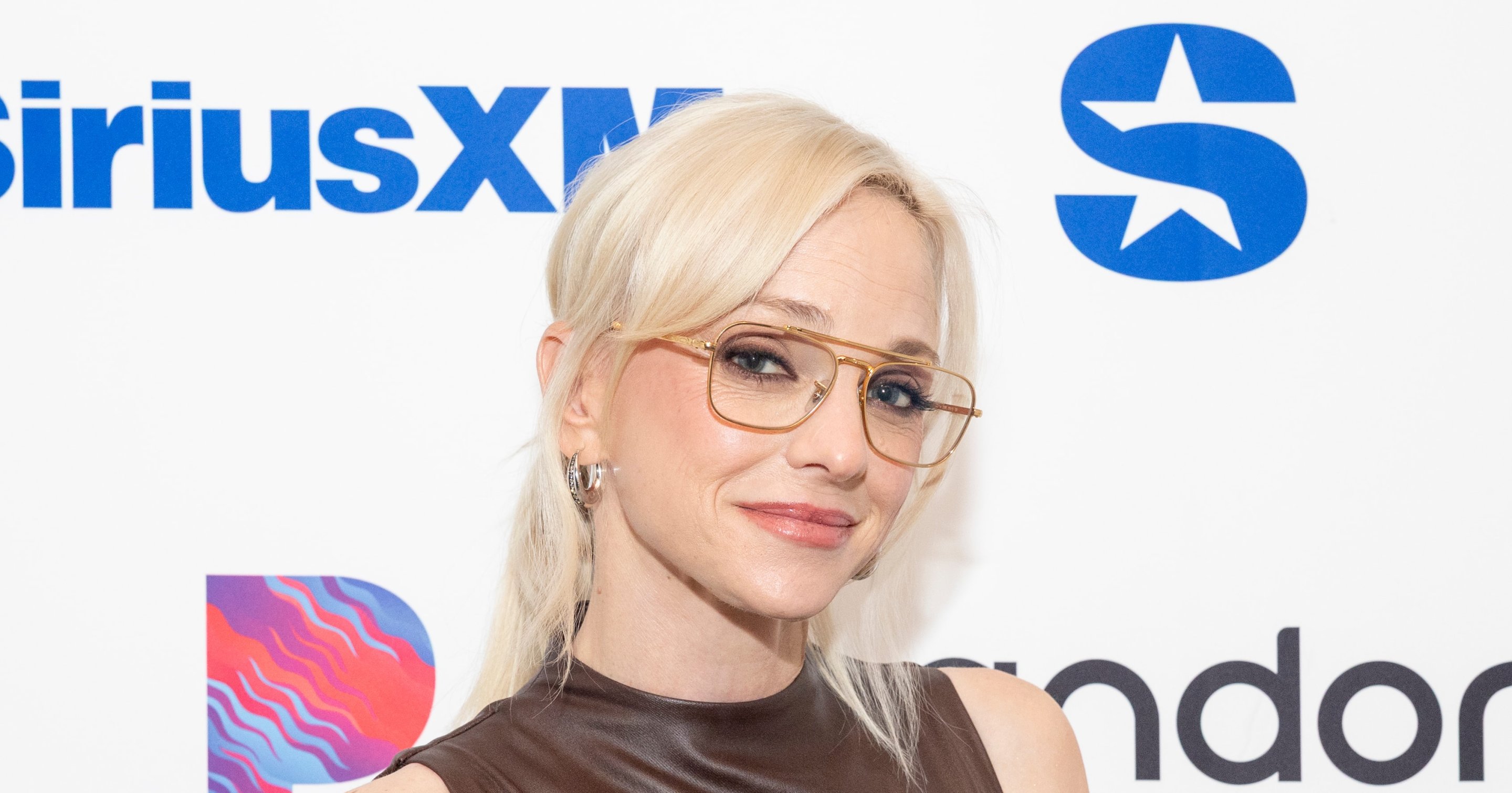 Anna Faris wearing glasses and a brown leather sleeveless top, smiling in front of a SiriusXM and Pandora branded backdrop.