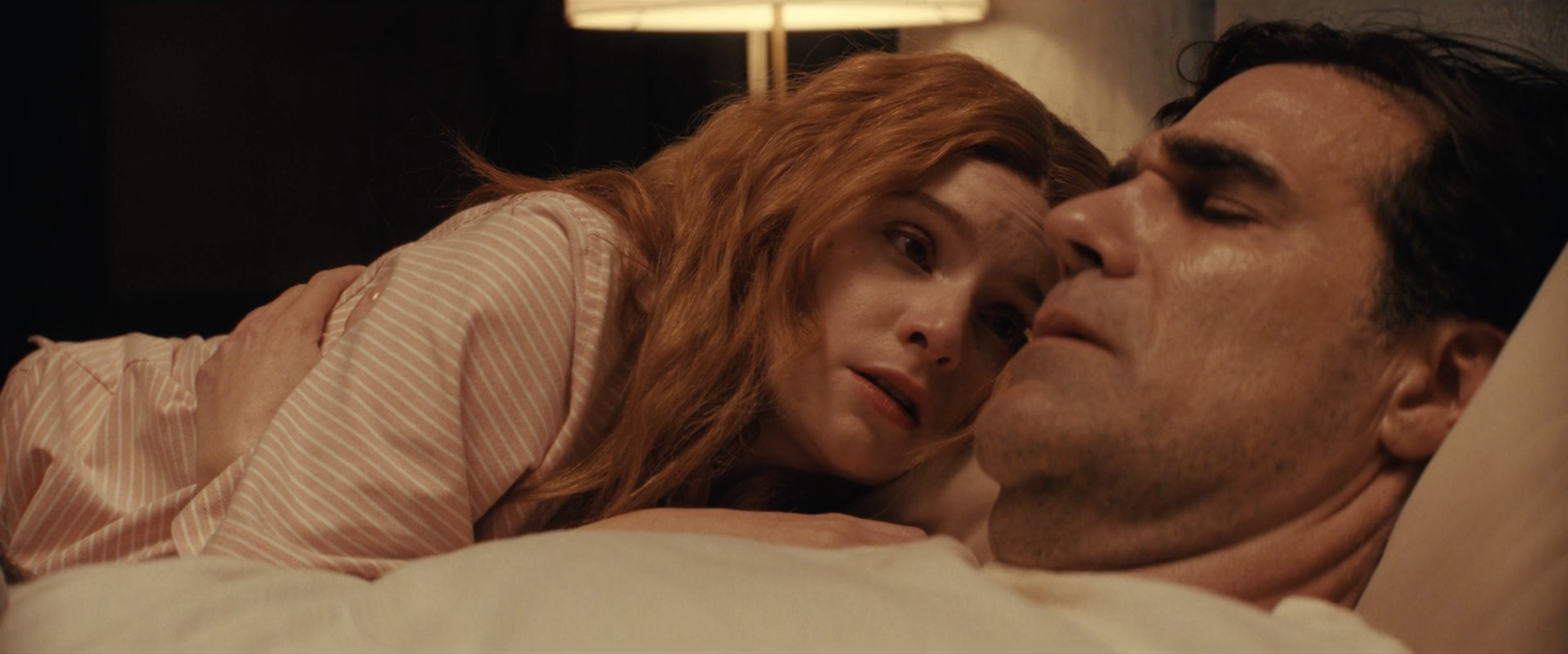 A red-haired woman in a striped shirt rests her head against a dark-haired man lying in bed, both lit by warm, low light.