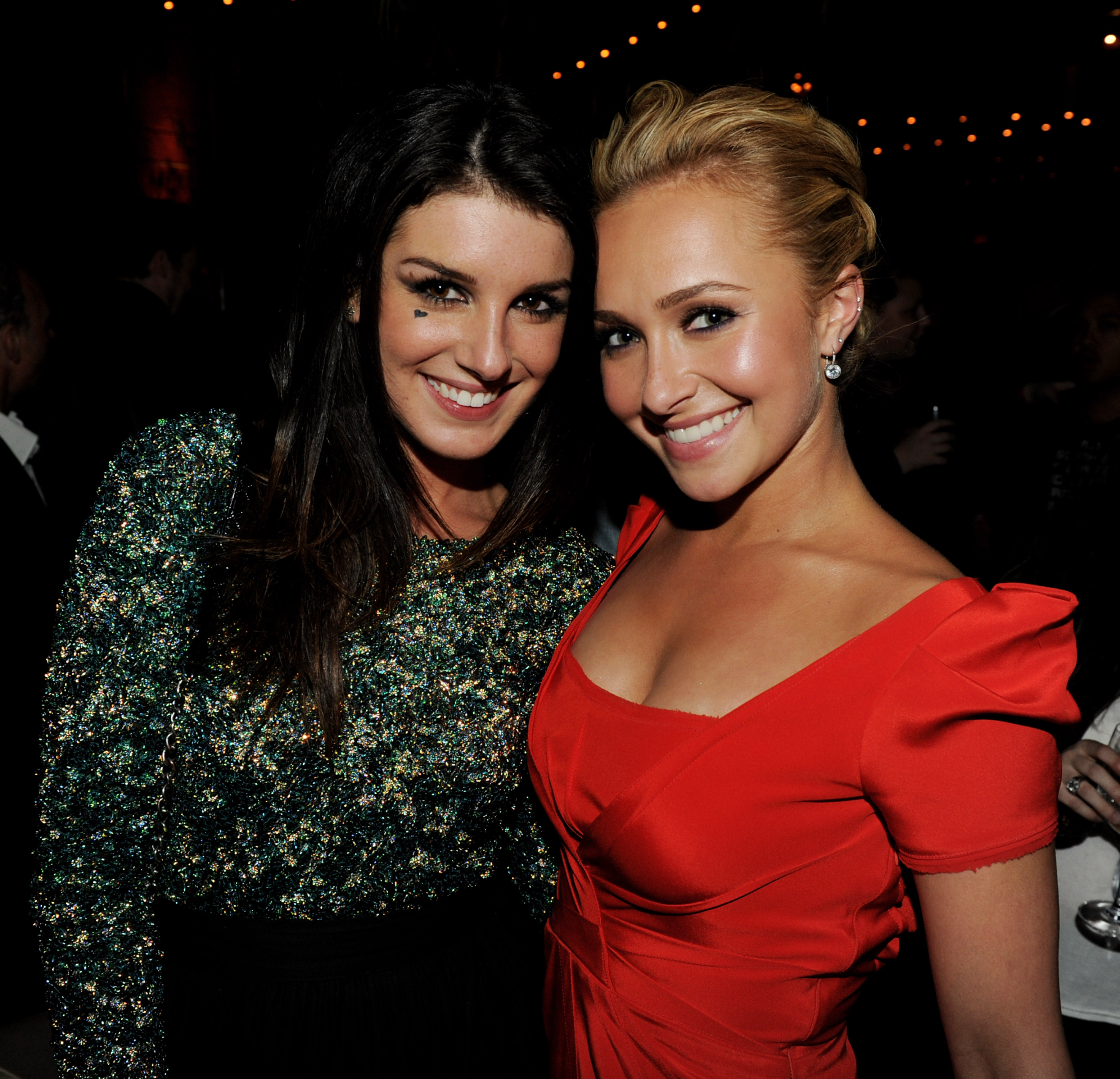 Two women smiling cheek to cheek at an indoor event, one in a dark glittery top and one in a red dress, with string lights in the background.