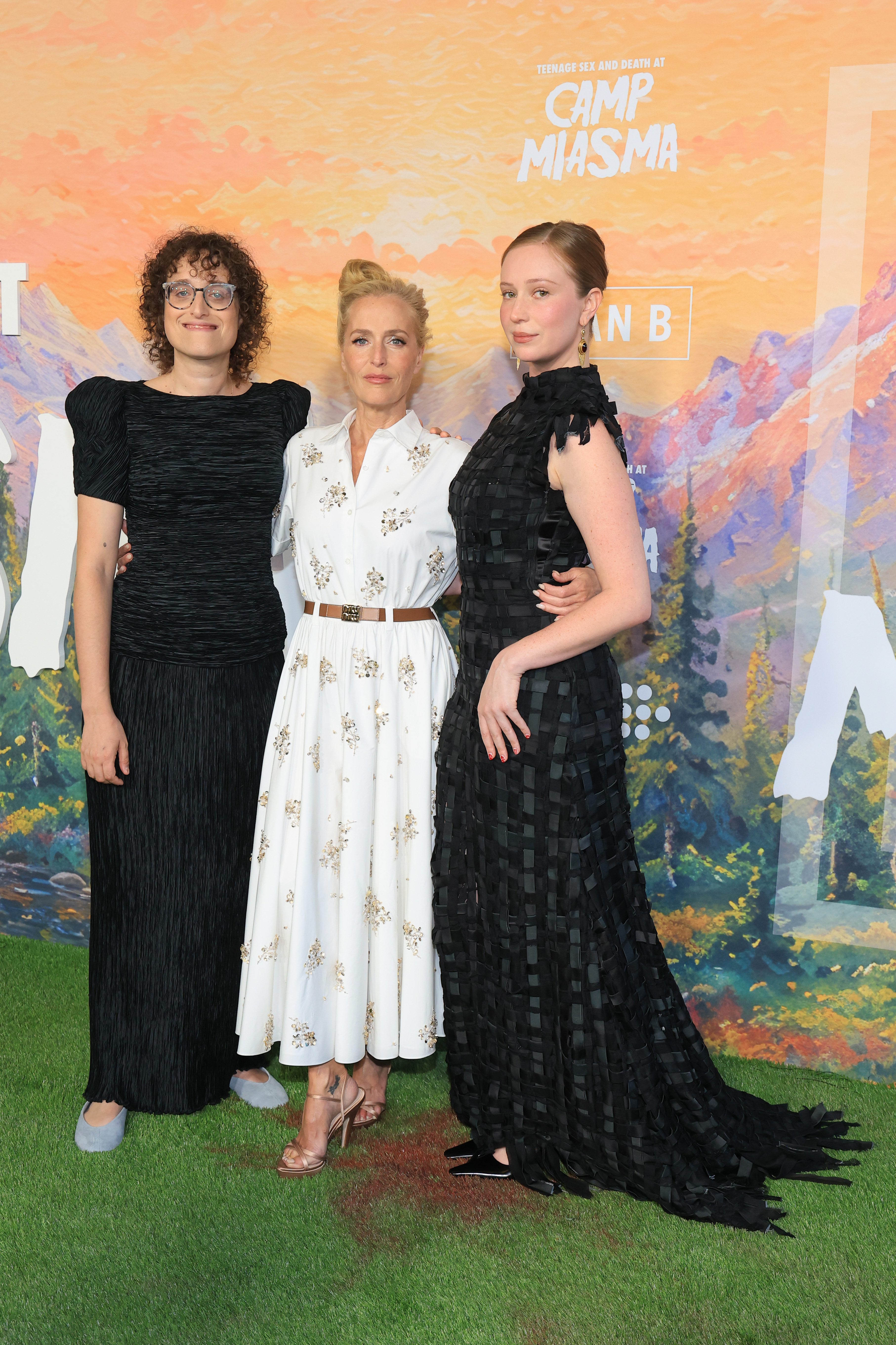 Jane Schoenbrun, Gillian Anderson, and Hannah Einbinder standing together in front of a Camp Miasma premiere backdrop at the DGA Theater Complex in Los Angeles.