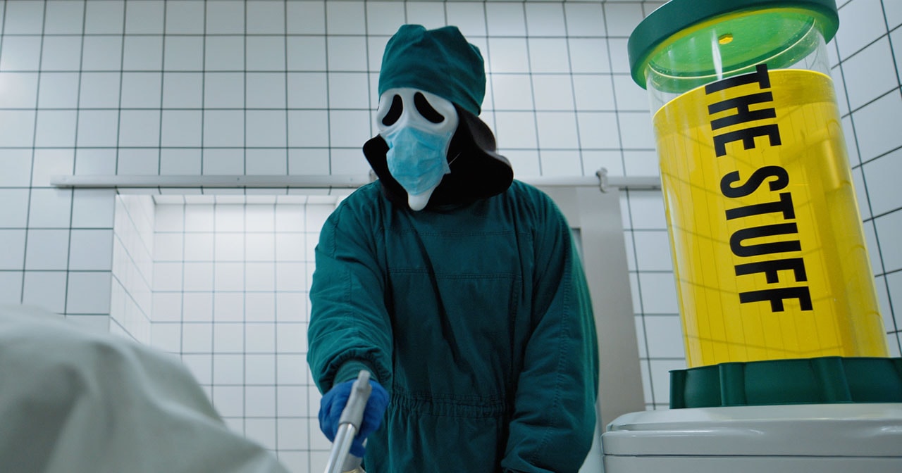 Person in Ghostface mask wearing teal surgical scrubs and a blue face mask stands in a tiled room next to a yellow canister labeled 'The Stuff.'