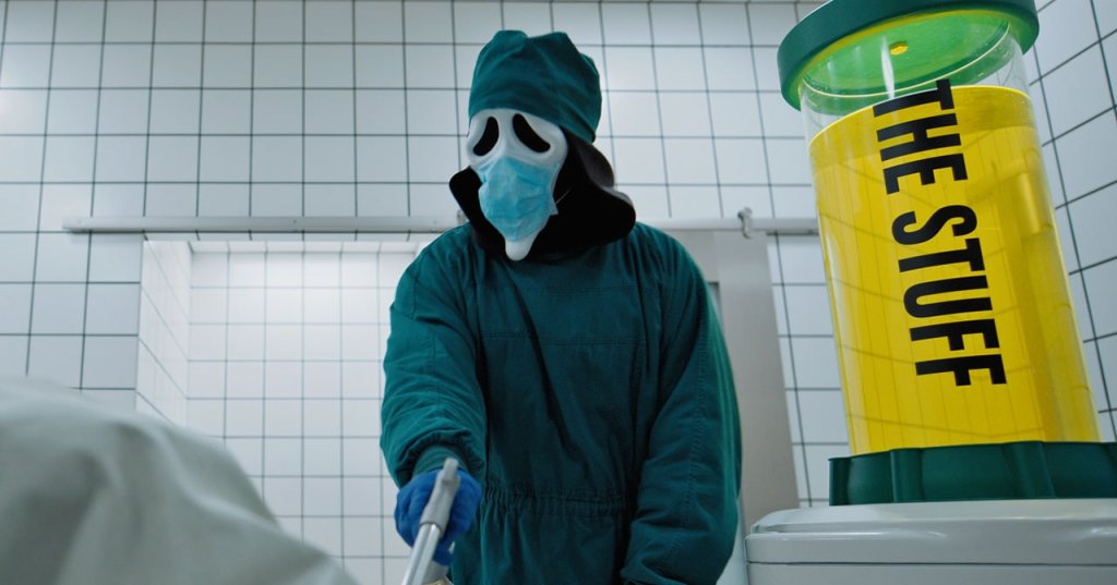 Person in Ghostface mask wearing teal surgical scrubs and a blue face mask stands in a tiled room next to a yellow canister labeled 'The Stuff.'