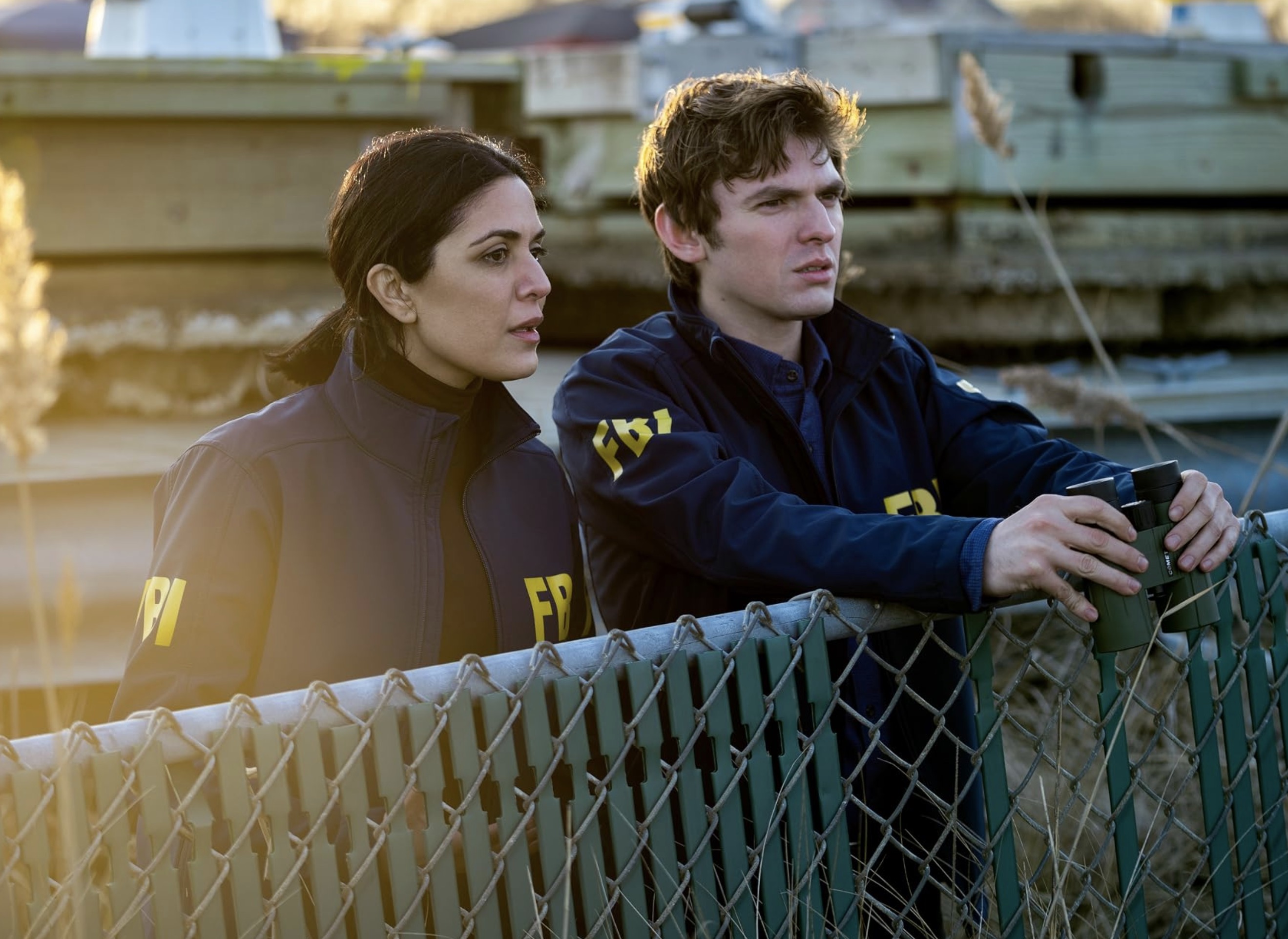 Two actors in FBI jackets lean over a chain-link fence, one holding binoculars, with industrial structures in the background.