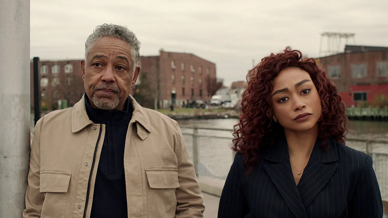 A middle-aged man in a beige jacket and a young woman with red curly hair stand together near a waterfront with brick buildings behind them.
