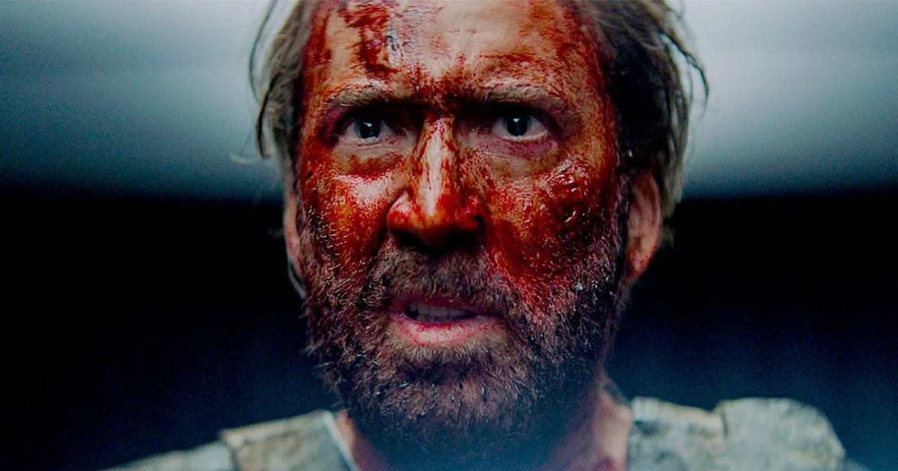 Close-up of a man with a beard, his face heavily covered in blood, staring intensely at the camera against a dark background.