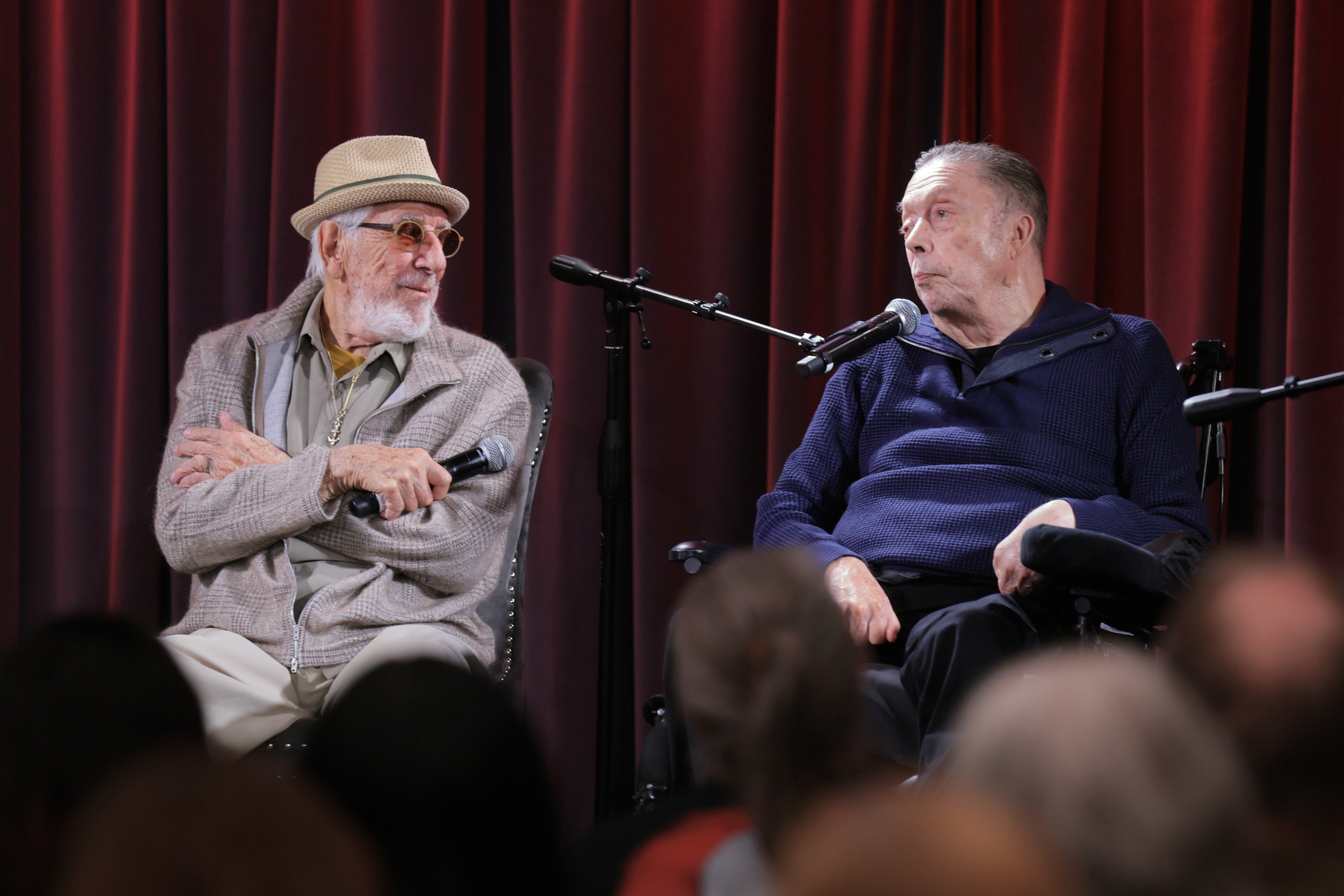 Two elderly men seated on a stage in front of red curtains, one wearing a straw hat and holding a microphone, the other in a blue sweater speaking into a mic stand.