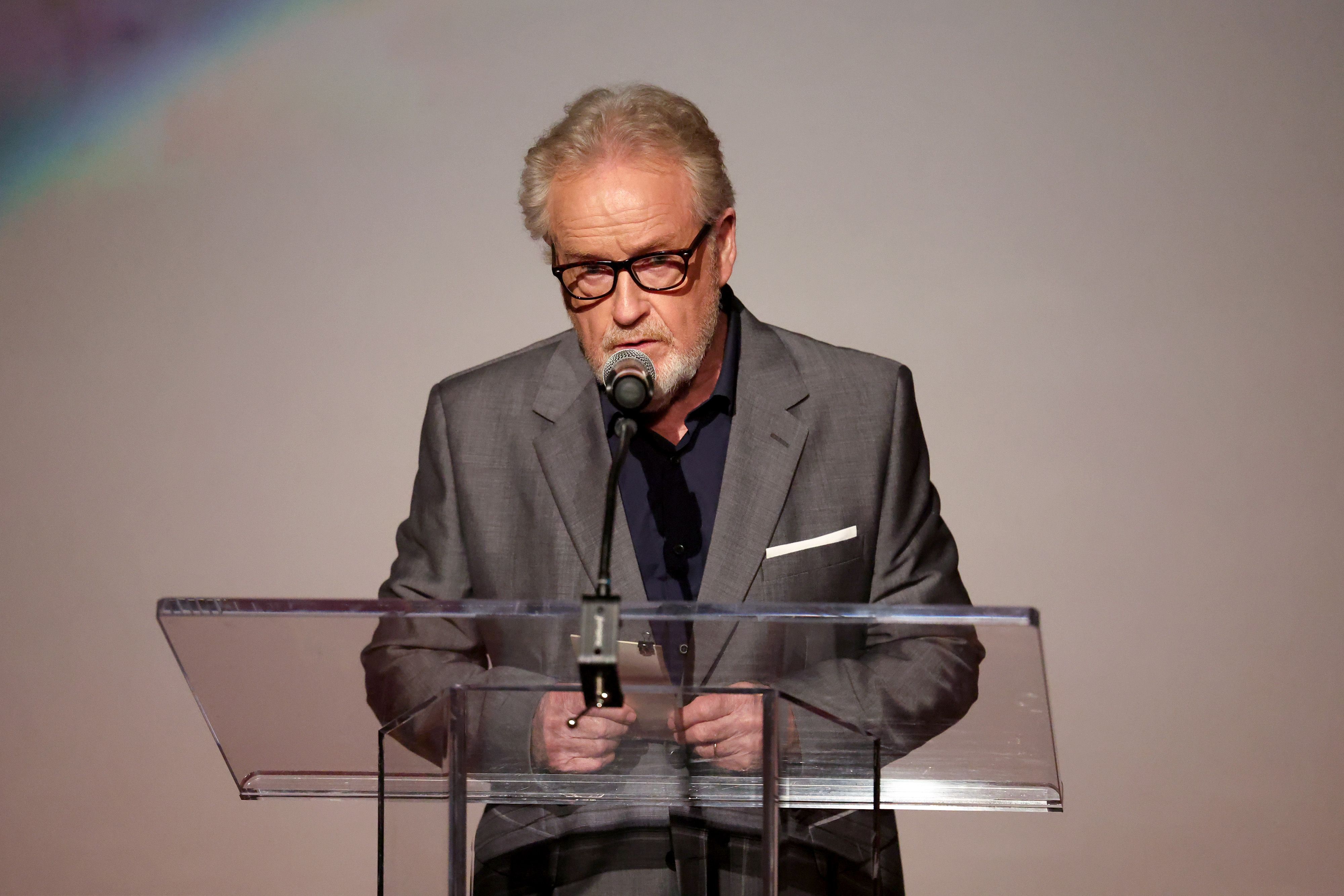Man in a gray blazer and glasses speaking into a microphone at a clear acrylic lectern on a stage.