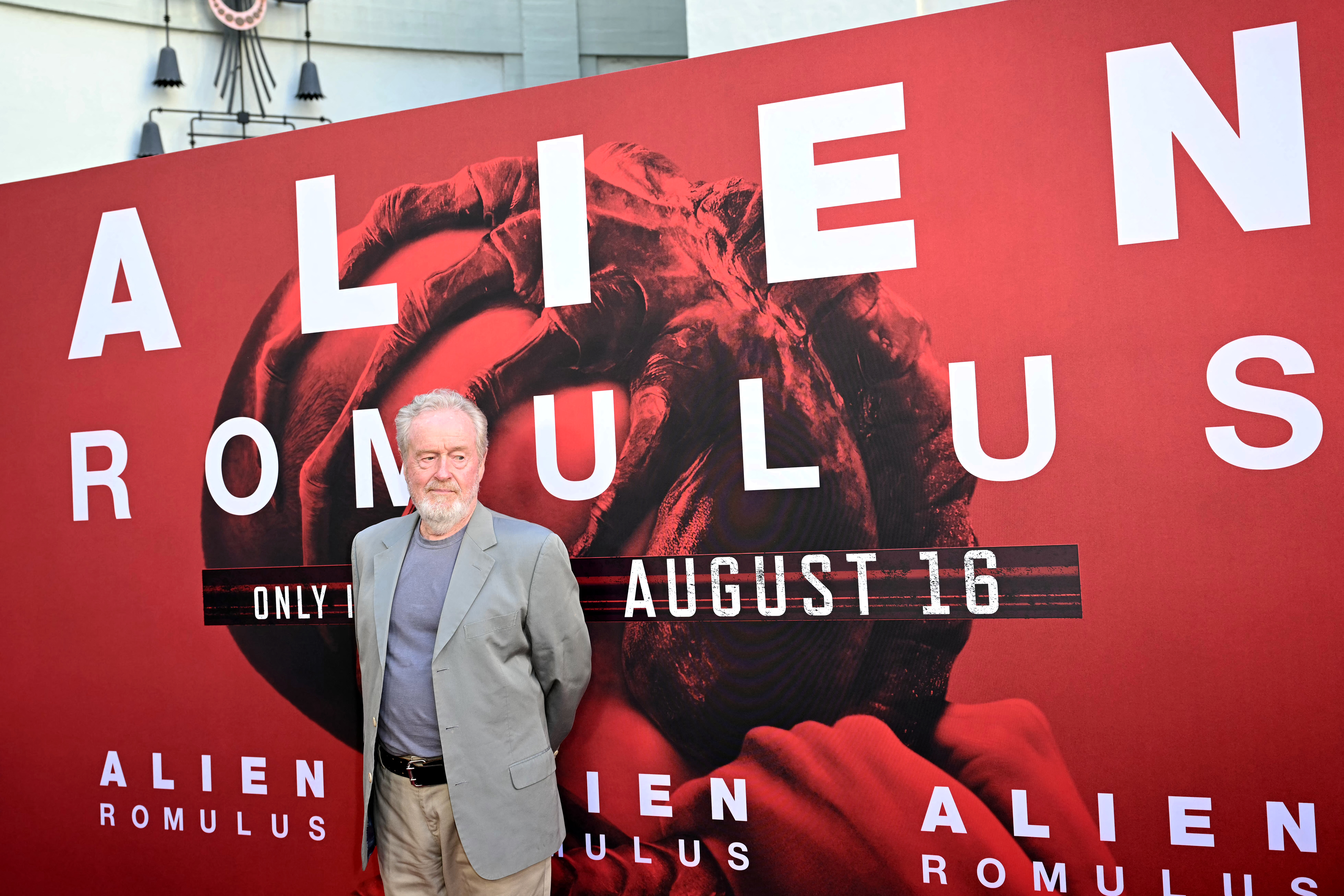 Man in a gray blazer and khakis standing before a large red Alien: Romulus movie poster announcing an August 16 release.