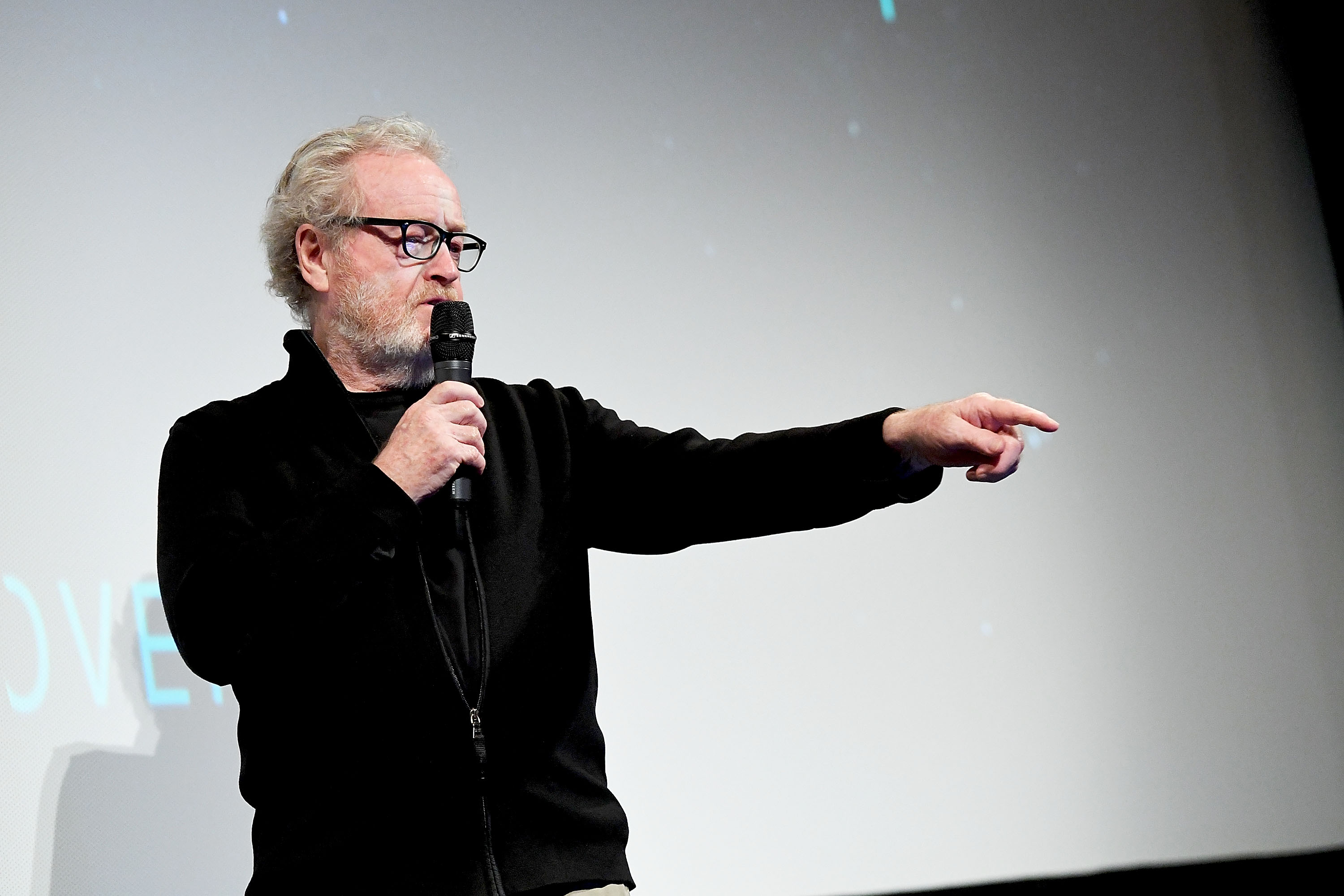 Older man in black wearing glasses holds a microphone and points into the audience in front of a large projection screen.