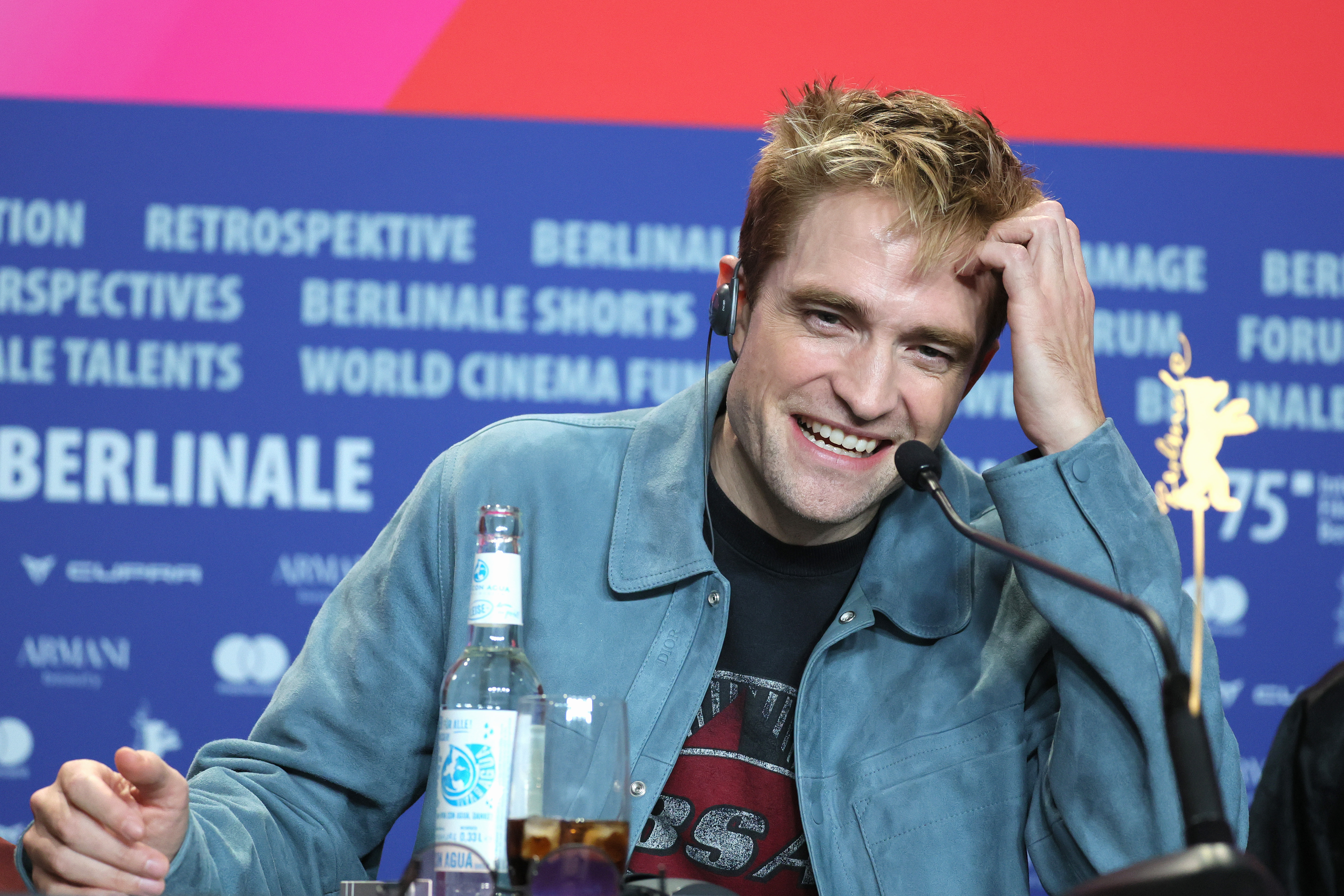 Man in blue-grey jacket laughing at a press conference table with a microphone, water bottle, and a Golden Bear award in front of a Berlinale banner.
