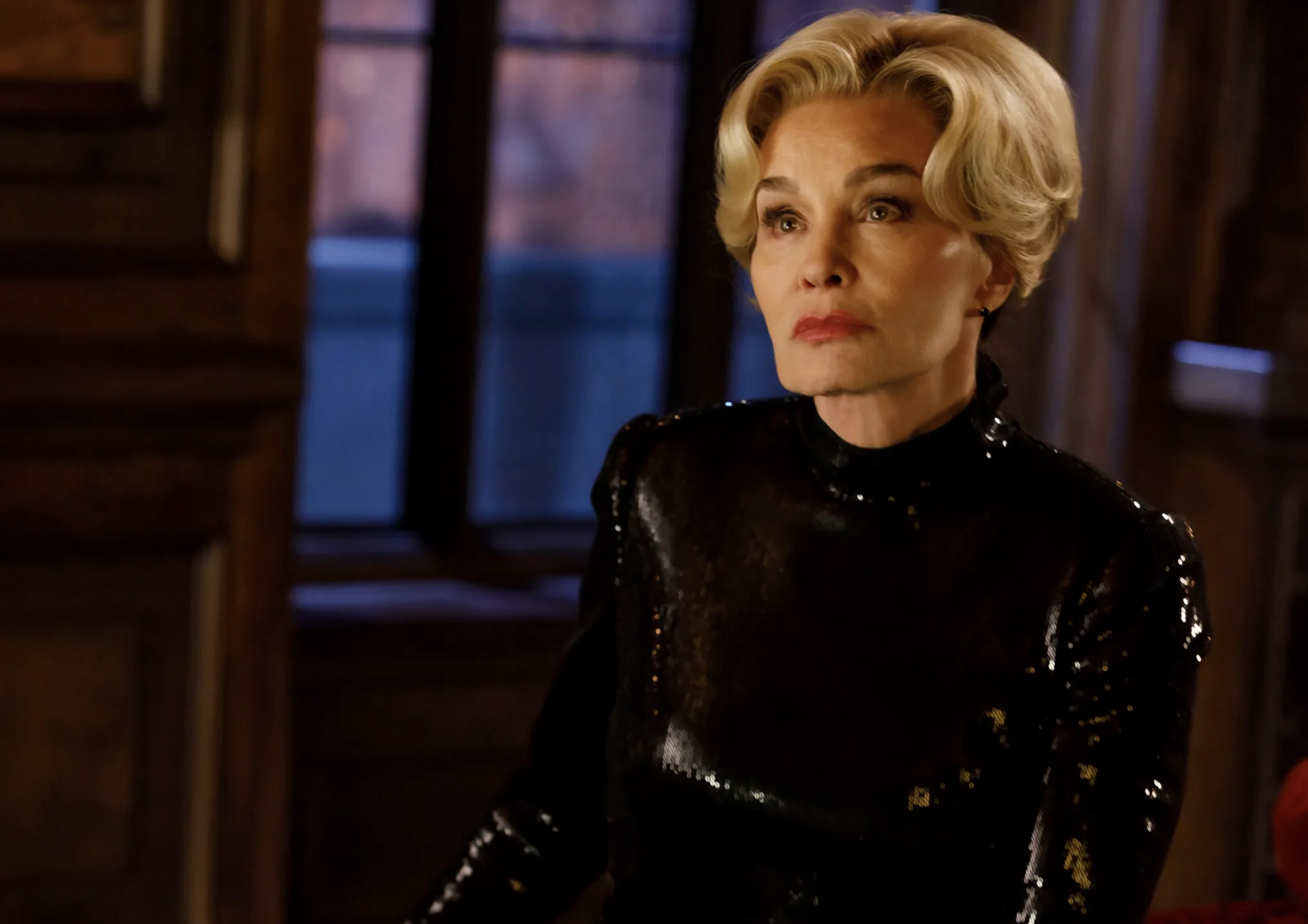 Woman with short blonde hair wearing a black sequined turtleneck standing in a dark wood-paneled room.