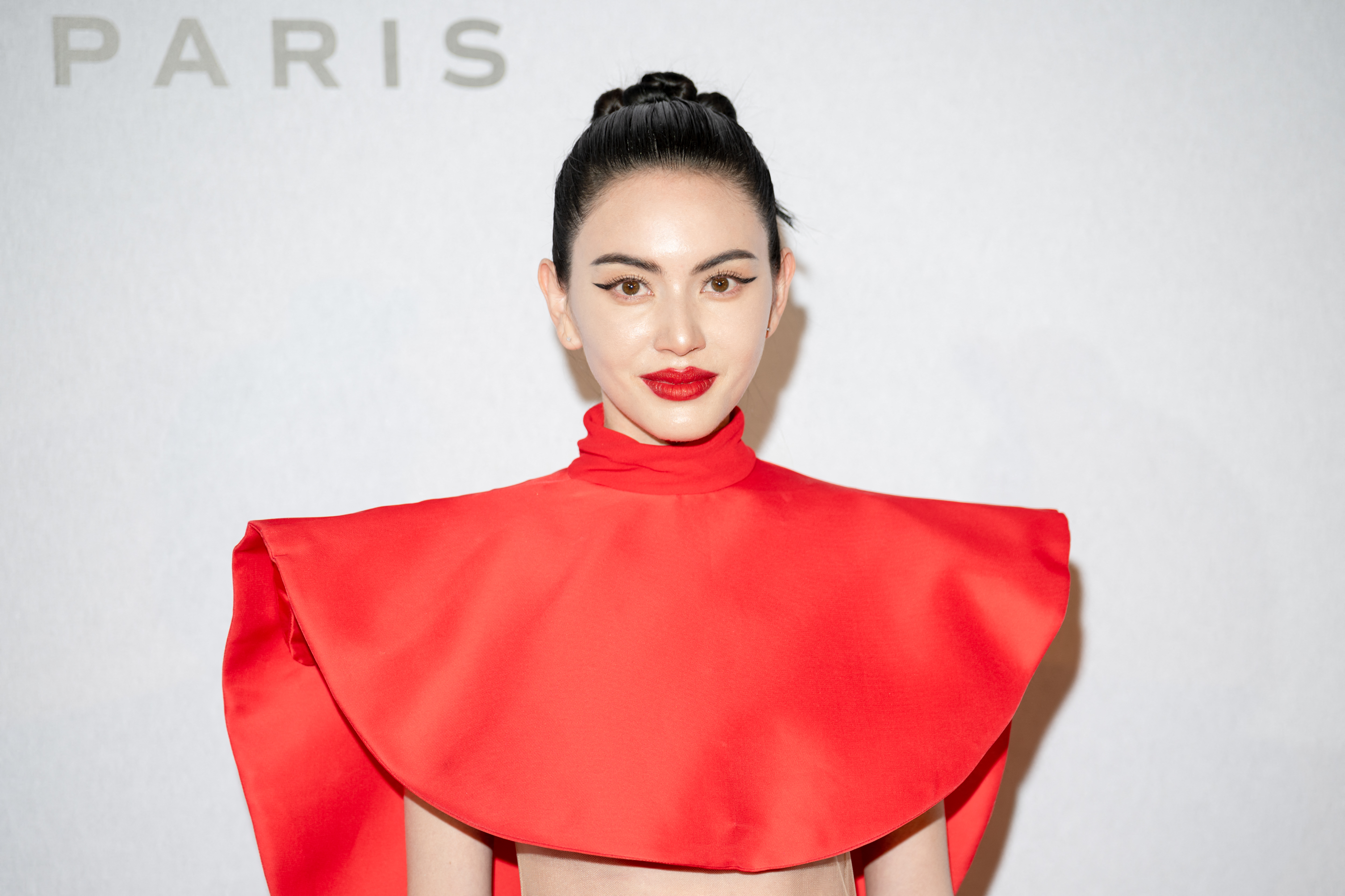 Woman in a bold red high-neck sculptural gown with wide collar, dark hair in a bun, red lips, at a Paris Fashion Week photocall backdrop.