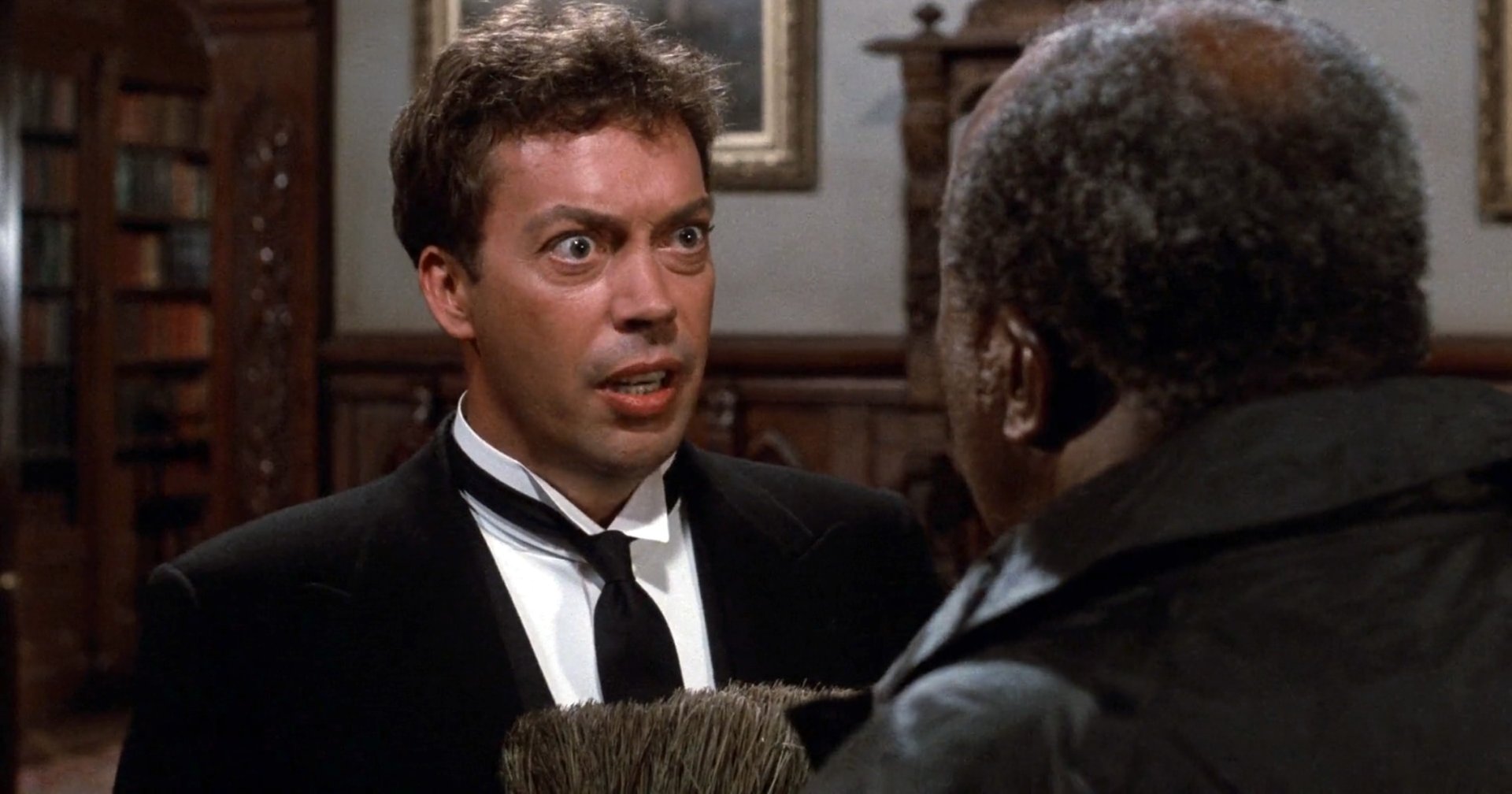 A wide-eyed man in a black tuxedo with a loosened tie faces another man in a wood-paneled room, mouth open mid-speech.