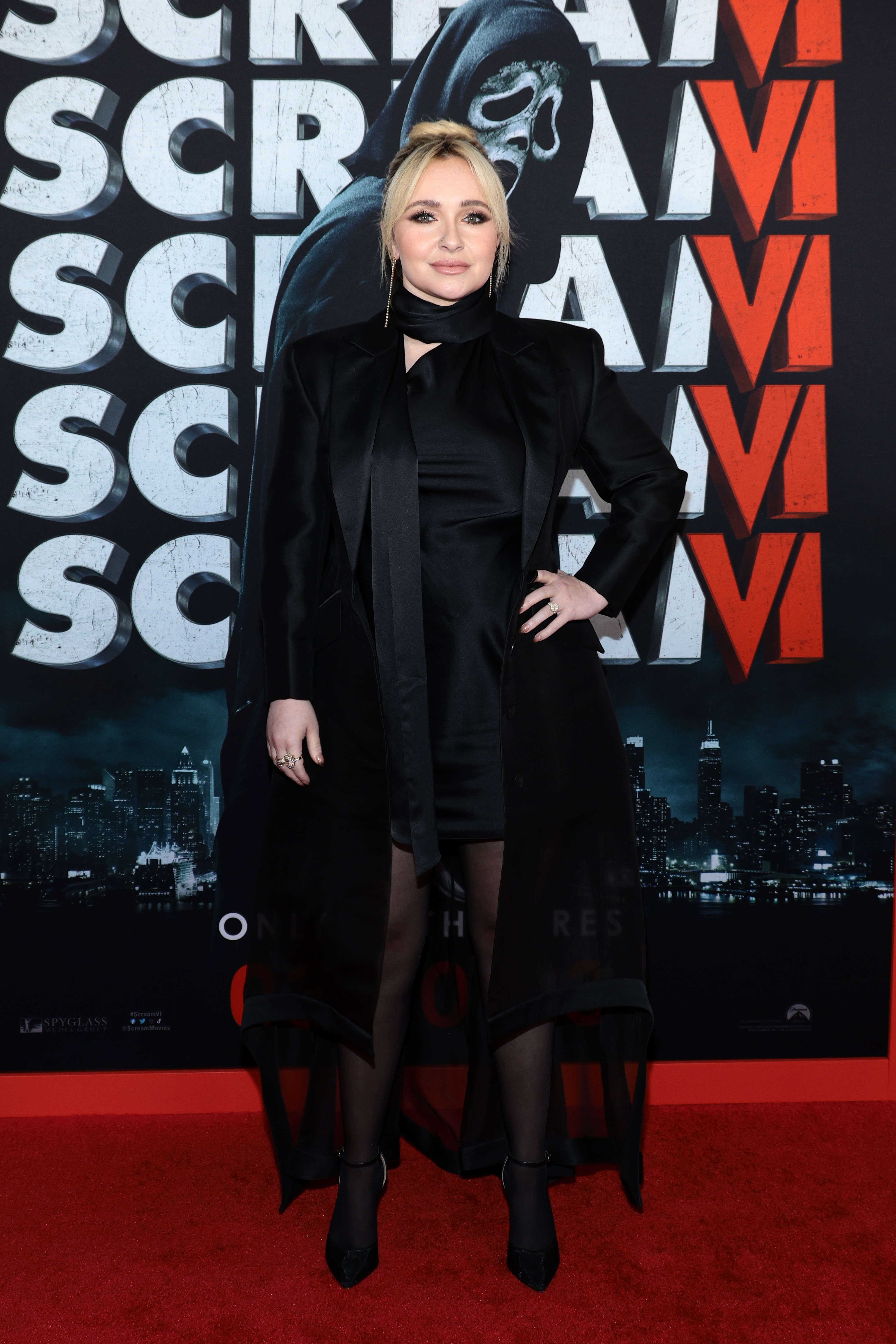 Hayden Panettiere in a black coat and dress posing in front of a Scream VI movie poster backdrop.