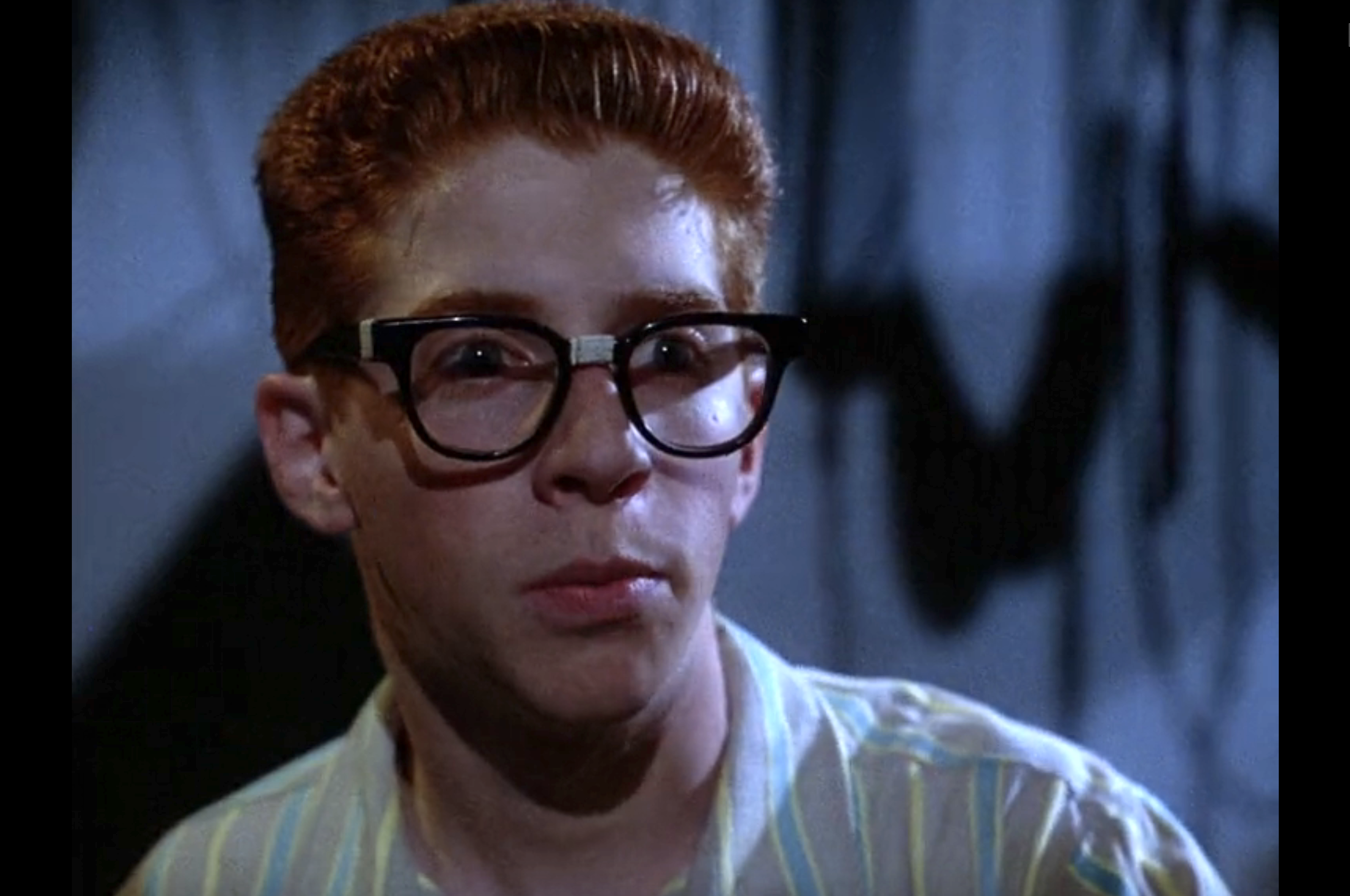Red-haired boy wearing large black-framed glasses and a striped shirt, looking fearful in a dimly lit scene.