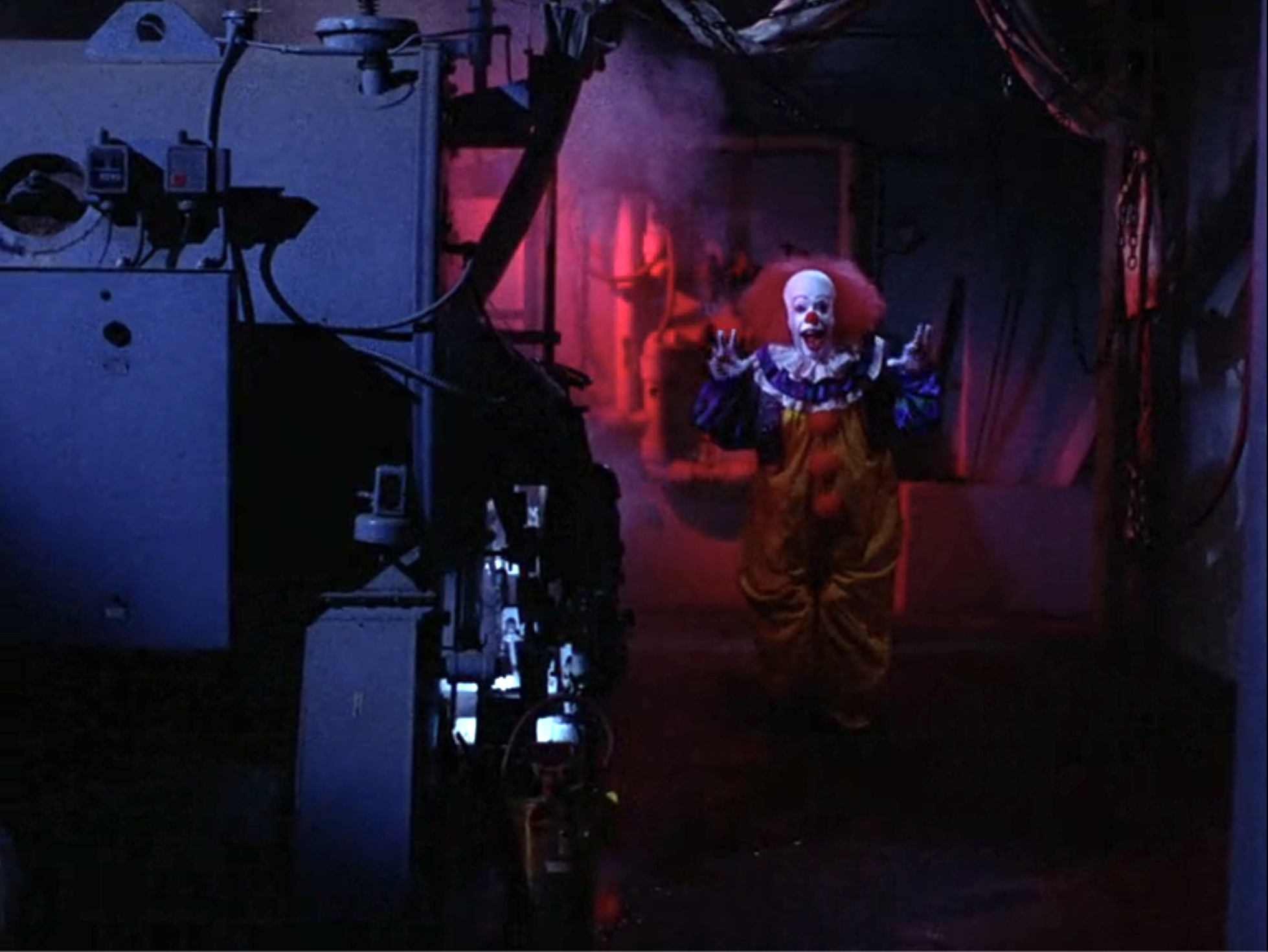 Pennywise the clown in yellow and purple costume standing in a dark, smoky industrial room with arms raised, bathed in red light.