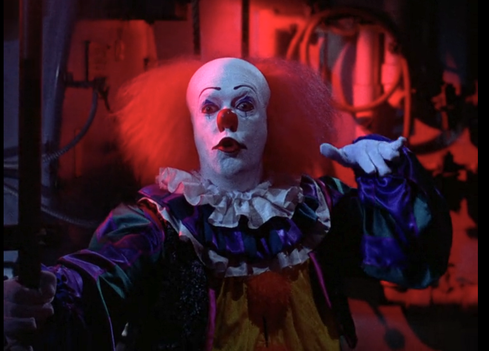 Pennywise the clown with white face paint, red hair, and colorful costume reaching arms forward in a dimly red-lit room.