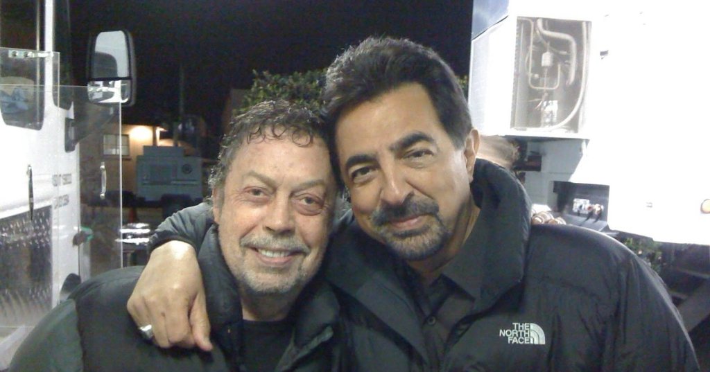 Two men in black puffer jackets smiling together on a nighttime film set, one with an arm around the other.