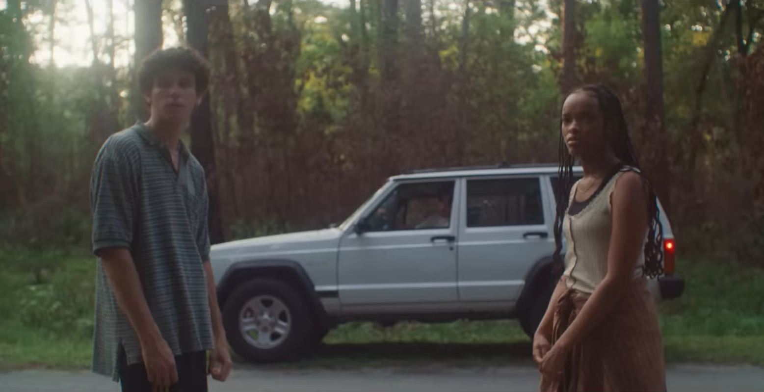 A teenage boy in a striped shirt and a girl with long braids stand apart on a wooded road with a white SUV parked behind them.
