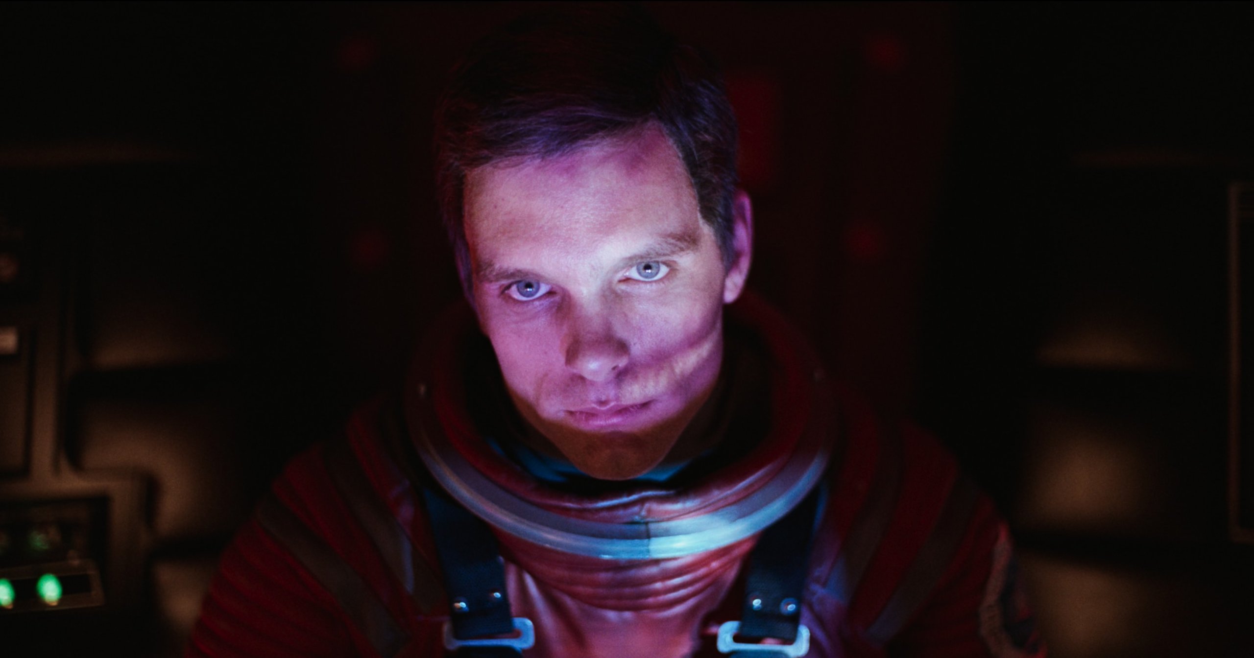 Close-up of an astronaut in a red space suit looking directly at the camera inside a dark spacecraft cockpit with glowing control panels in the background.