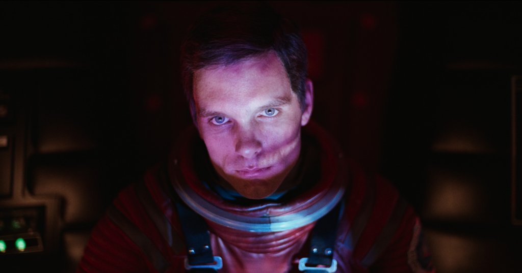 Close-up of an astronaut in a red space suit looking directly at the camera inside a dark spacecraft cockpit with glowing control panels in the background.