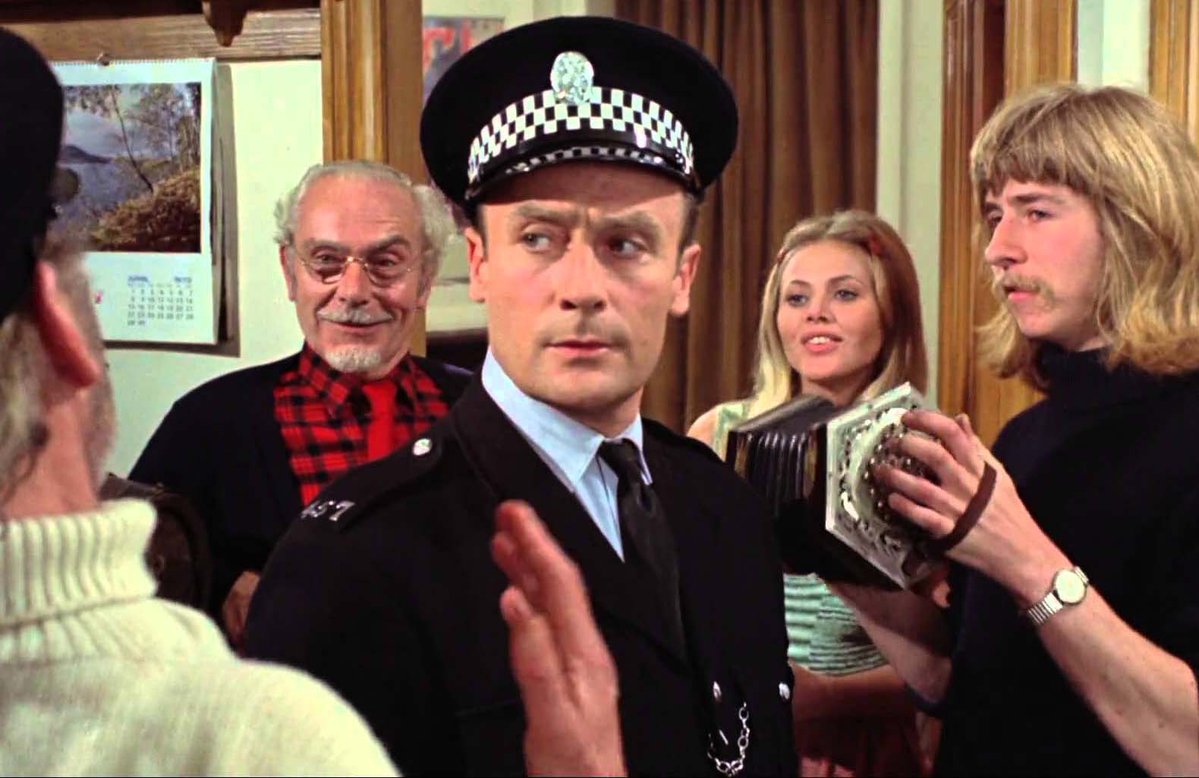 A man in a British police uniform and checkered cap stands amid a group of quirky locals, one of whom plays a concertina, in an indoor scene.