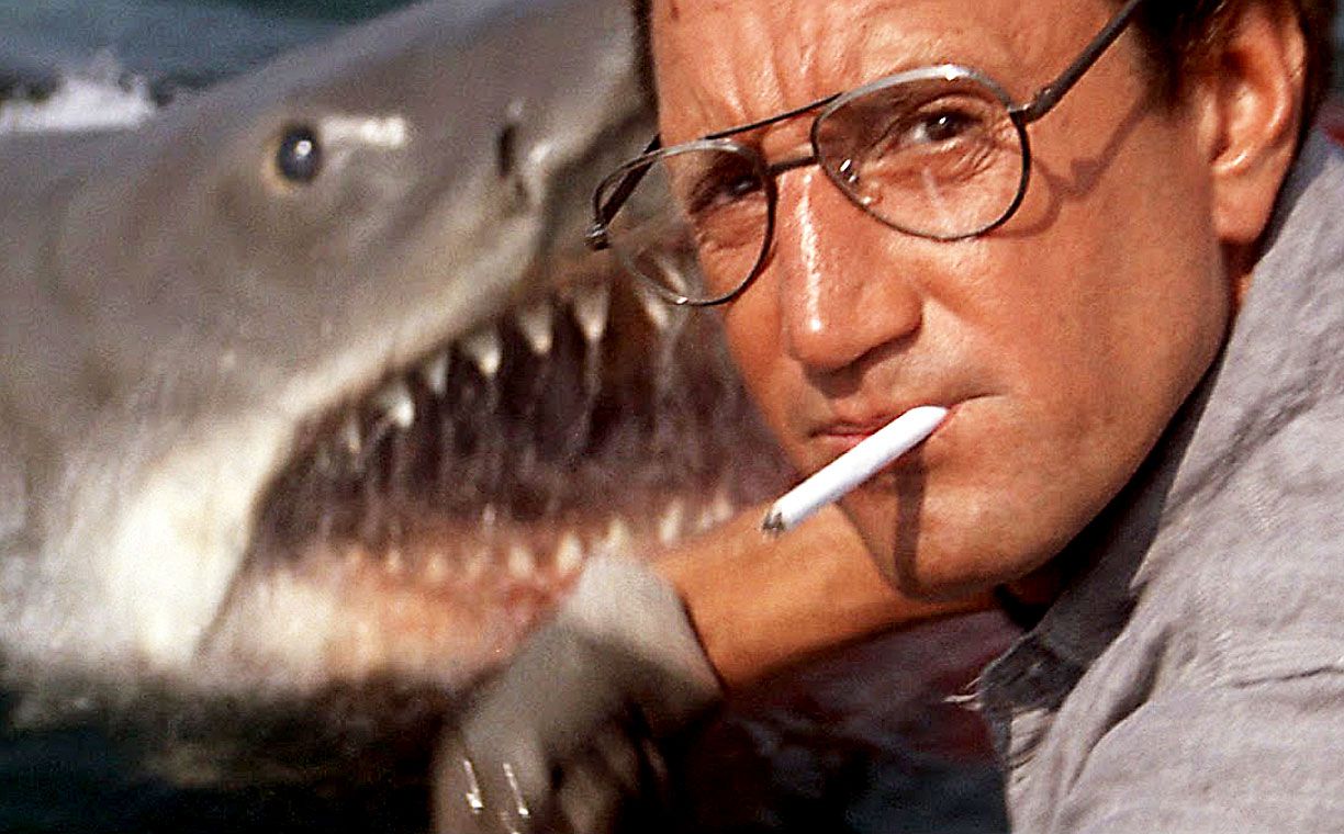 A middle-aged man with glasses and a cigarette stares ahead with a large mechanical shark's open-mouthed head looming behind him.