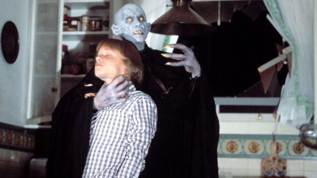 A blue-skinned vampire with yellow eyes and long fingernails grabs a boy in a plaid shirt by the throat in a kitchen.