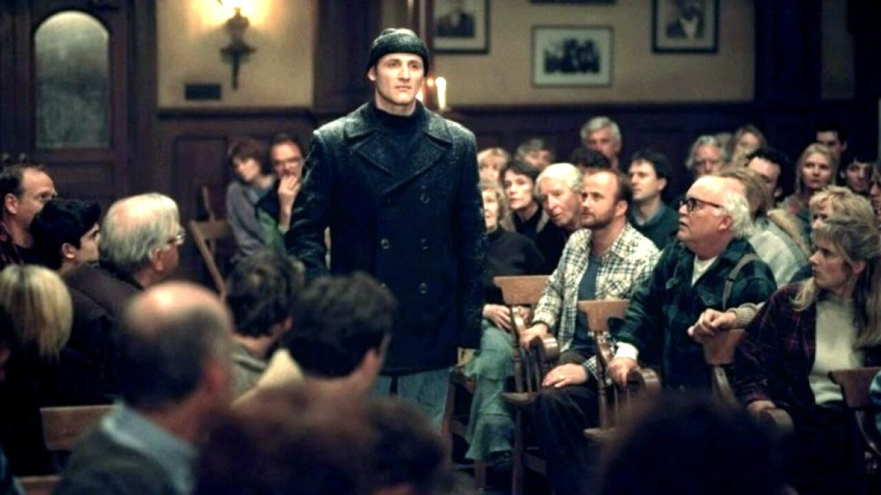 A tall man in a black coat and knit cap stands among rows of seated townspeople in a wood-paneled meeting hall.
