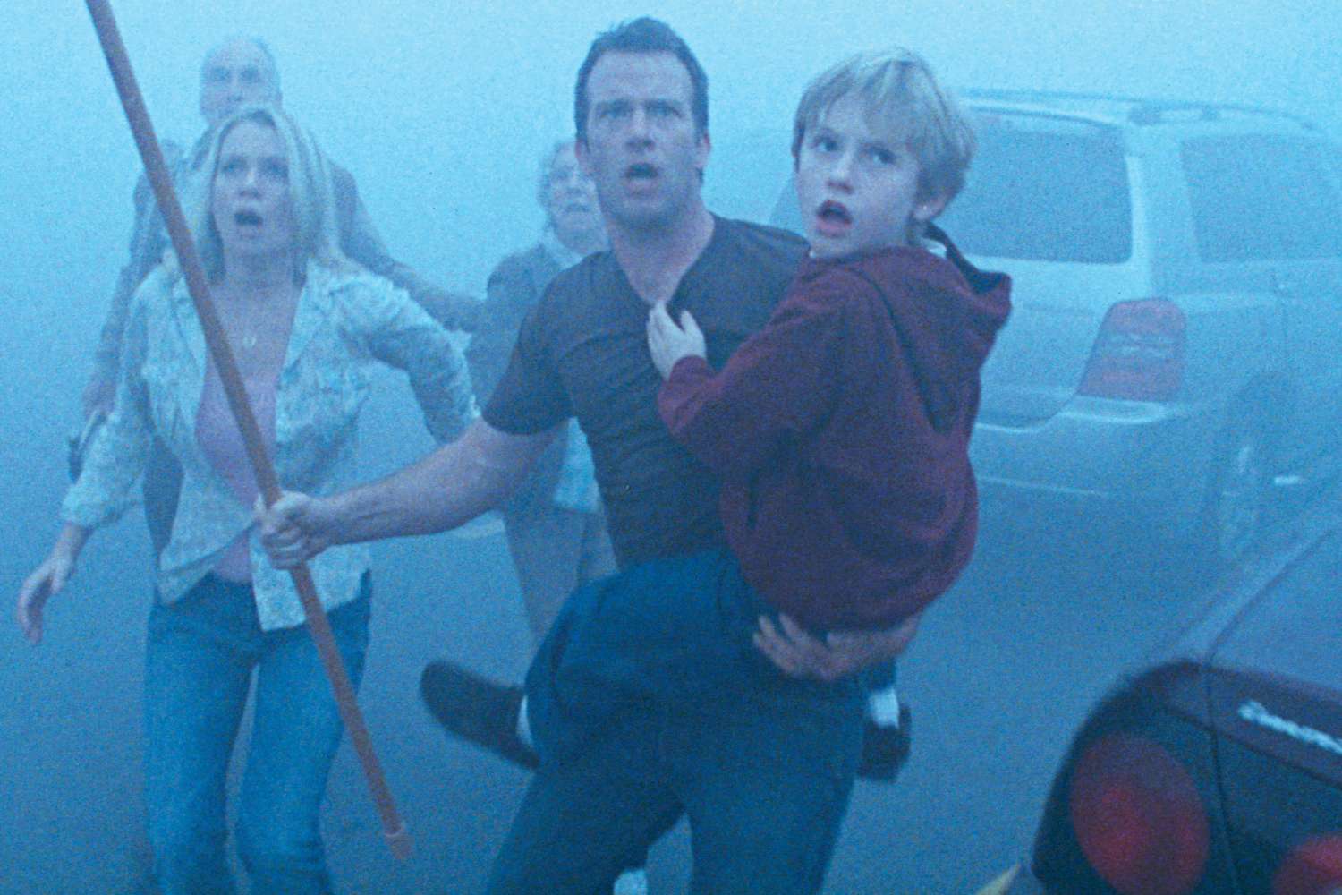 A woman holding a long stick and a man carrying a young boy run in fear through dense mist near parked cars.