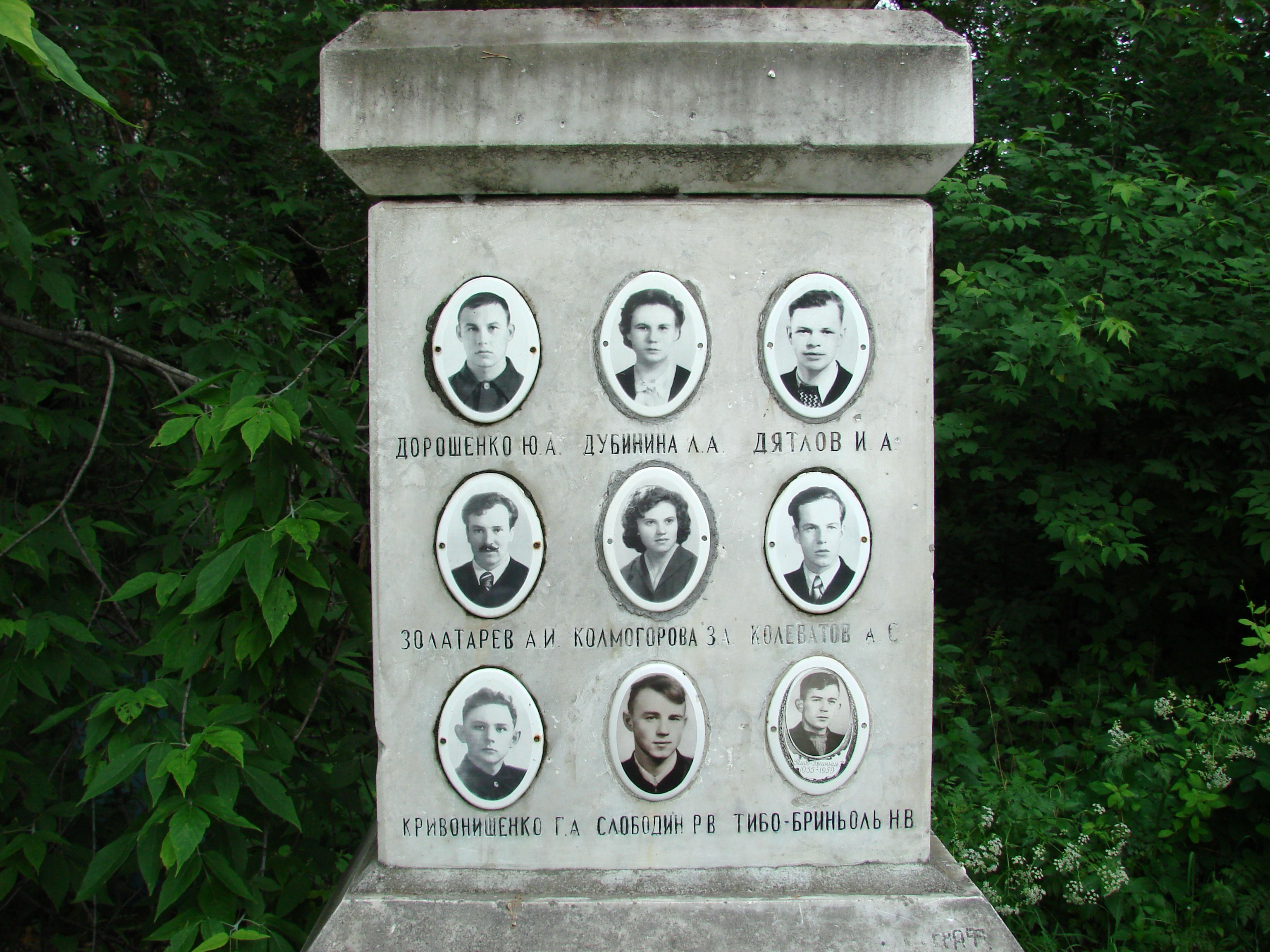 Stone memorial with nine oval black-and-white portrait photos and Cyrillic names of the Dyatlov Pass victims, set against green foliage.