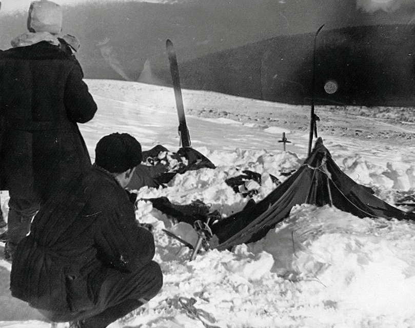 Black-and-white photo of two rescuers examining a collapsed, snow-buried tent on a mountain slope, with skis sticking upright from the snow nearby.