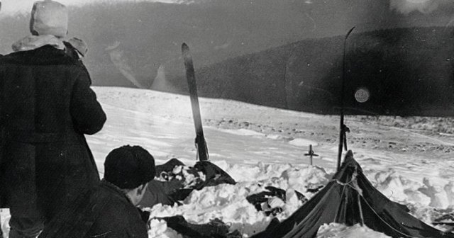 Black and white photo of two people crouching beside a collapsed, snow-covered tent with skis propped upright nearby on a snowy mountain slope.
