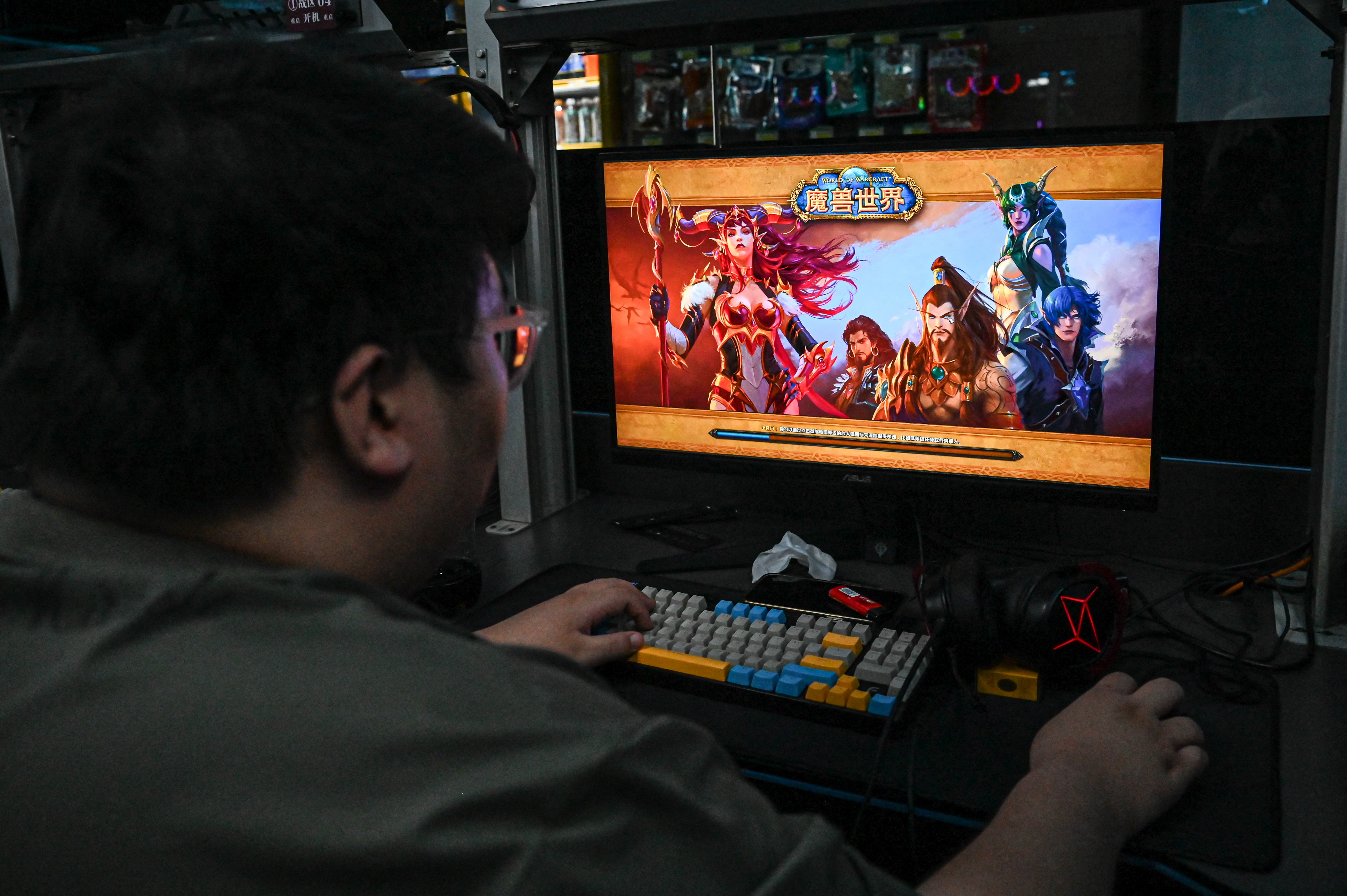A man seen from behind plays World of Warcraft on an ASUS monitor at a dimly lit internet cafe, with a colorful mechanical keyboard in front of him.