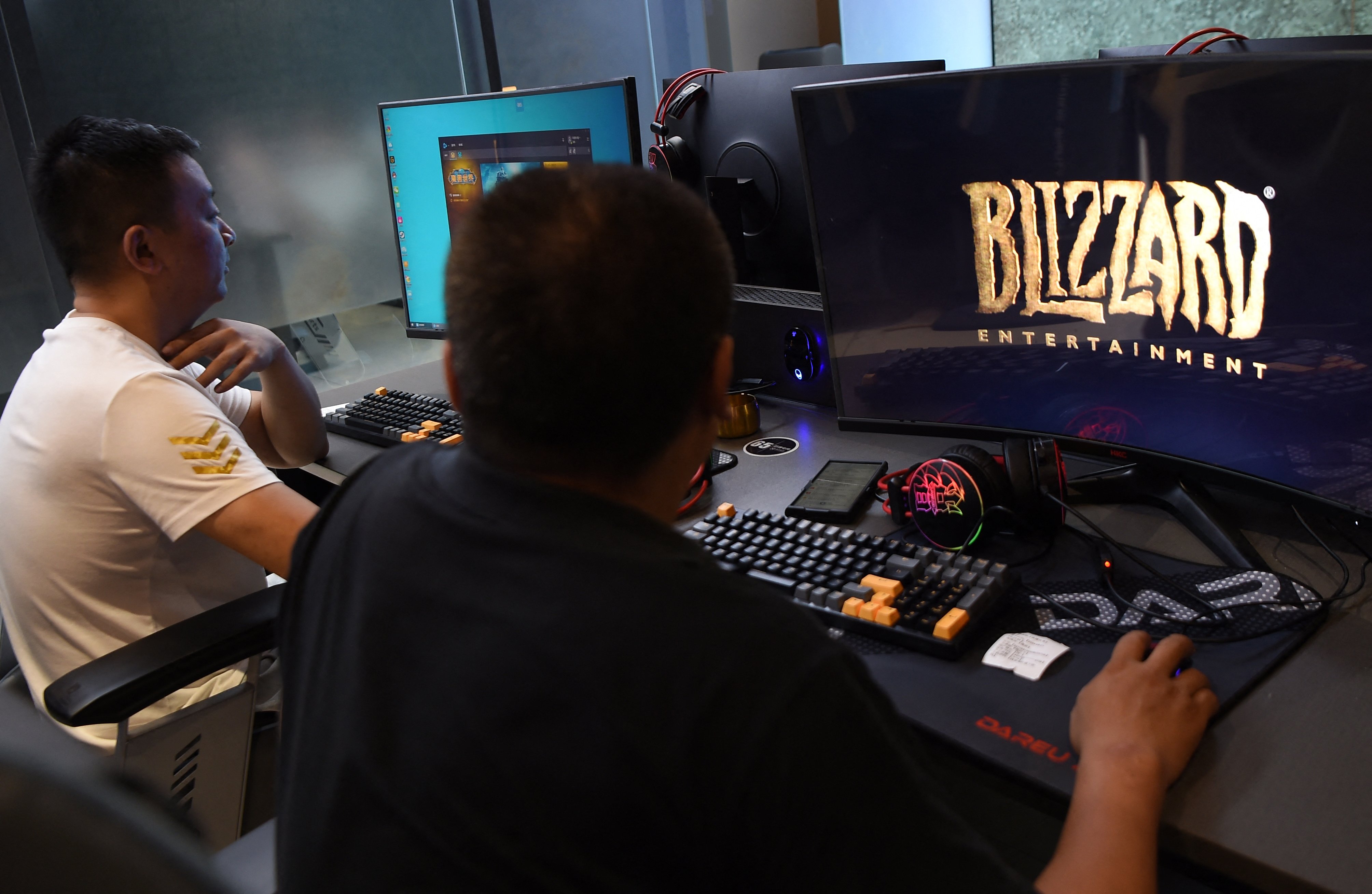 Two men at a gaming desk, one facing left and one seen from behind, with a large monitor showing the Blizzard Entertainment logo and a second monitor in the background.
