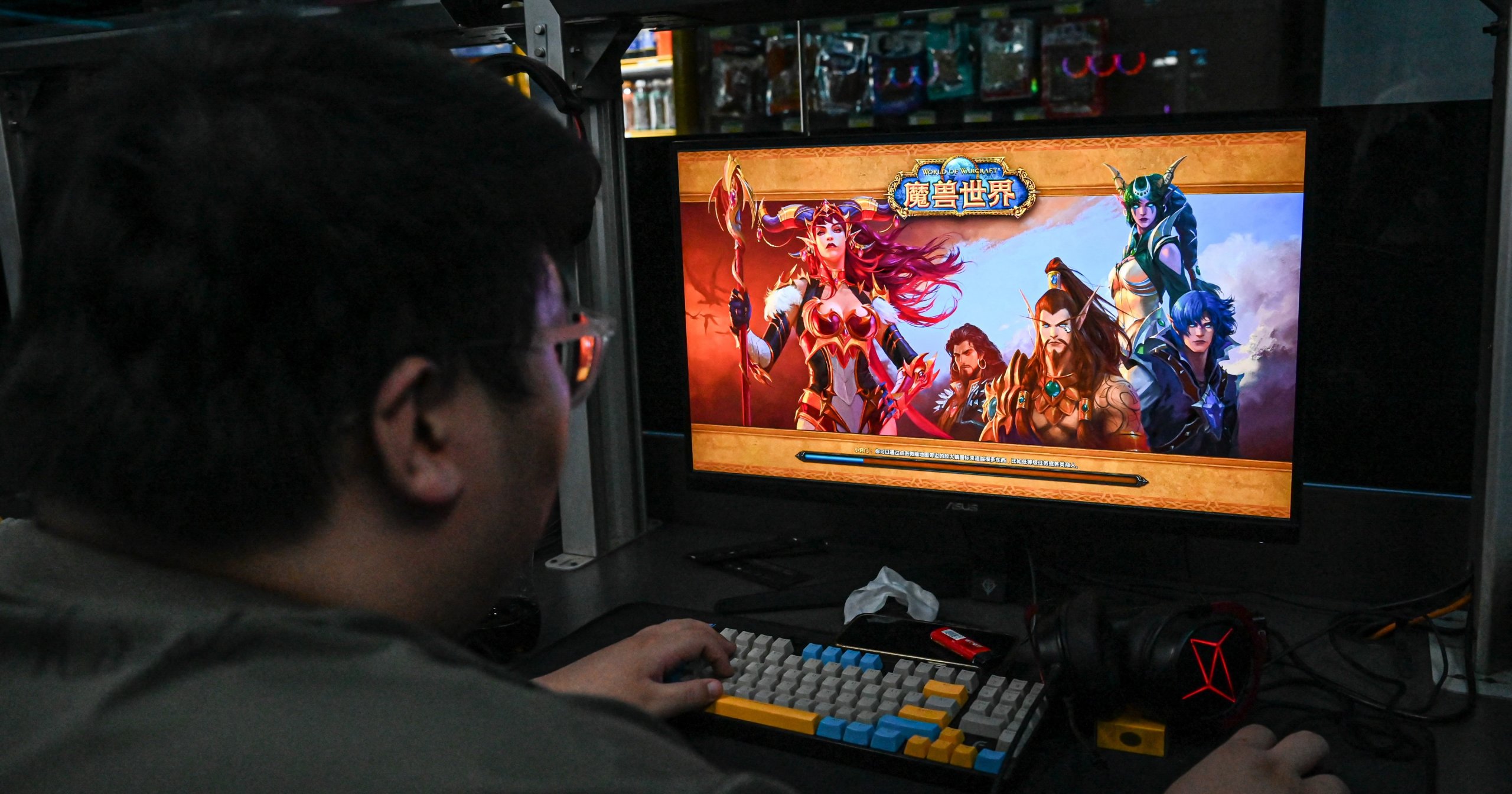 Man seen from behind playing World of Warcraft on a monitor at a dimly lit Beijing internet café, colorful keyboard and headset on desk.