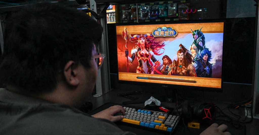 Man seen from behind playing World of Warcraft on a monitor at a dimly lit Beijing internet café, colorful keyboard and headset on desk.