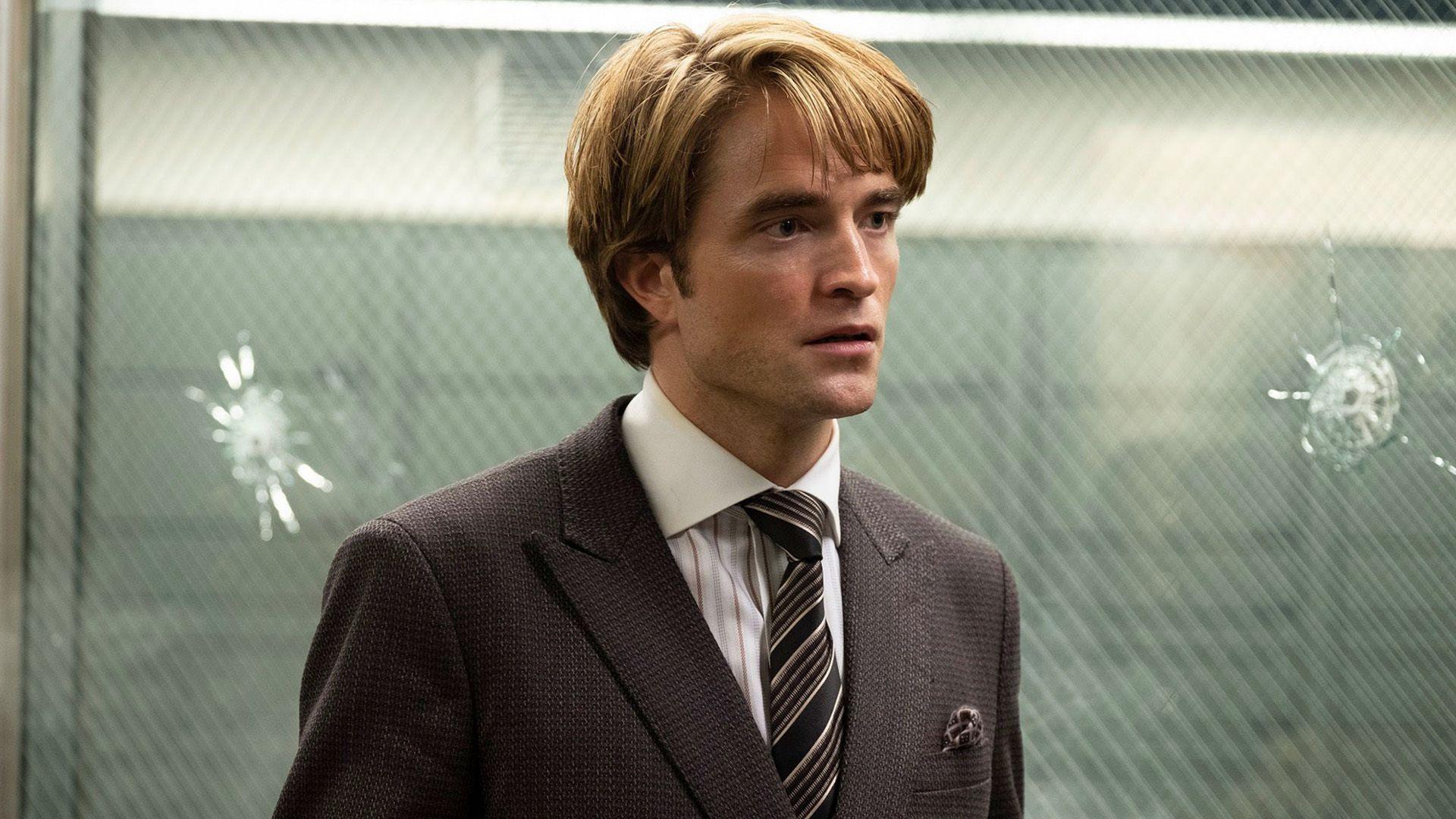 A young man with blond hair in a dark suit and striped tie stands before a glass panel with multiple bullet holes.