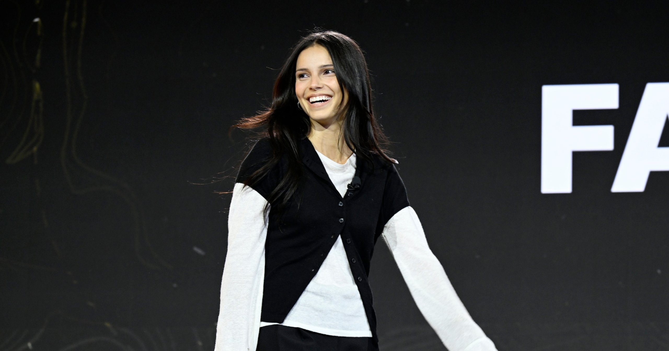 Actress Inde Navarrette smiling and gesturing onstage at Fanatics Fest NYC 2026, wearing a black cardigan over a white top.