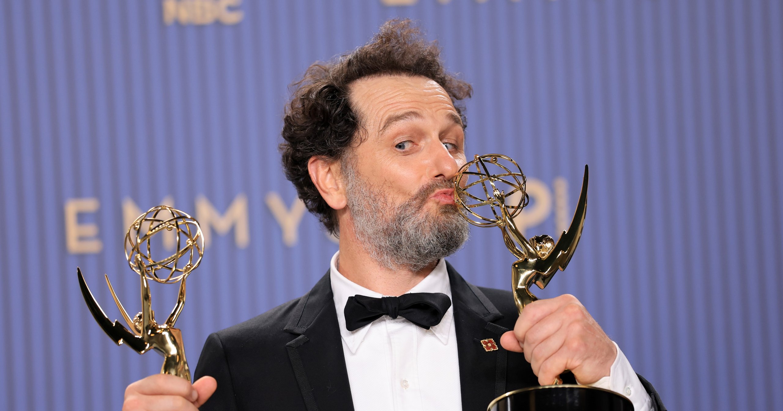 A bearded man in a tuxedo and bow tie kisses one of two gold Emmy Award statuettes he holds, grinning, in front of an Emmy-branded backdrop.