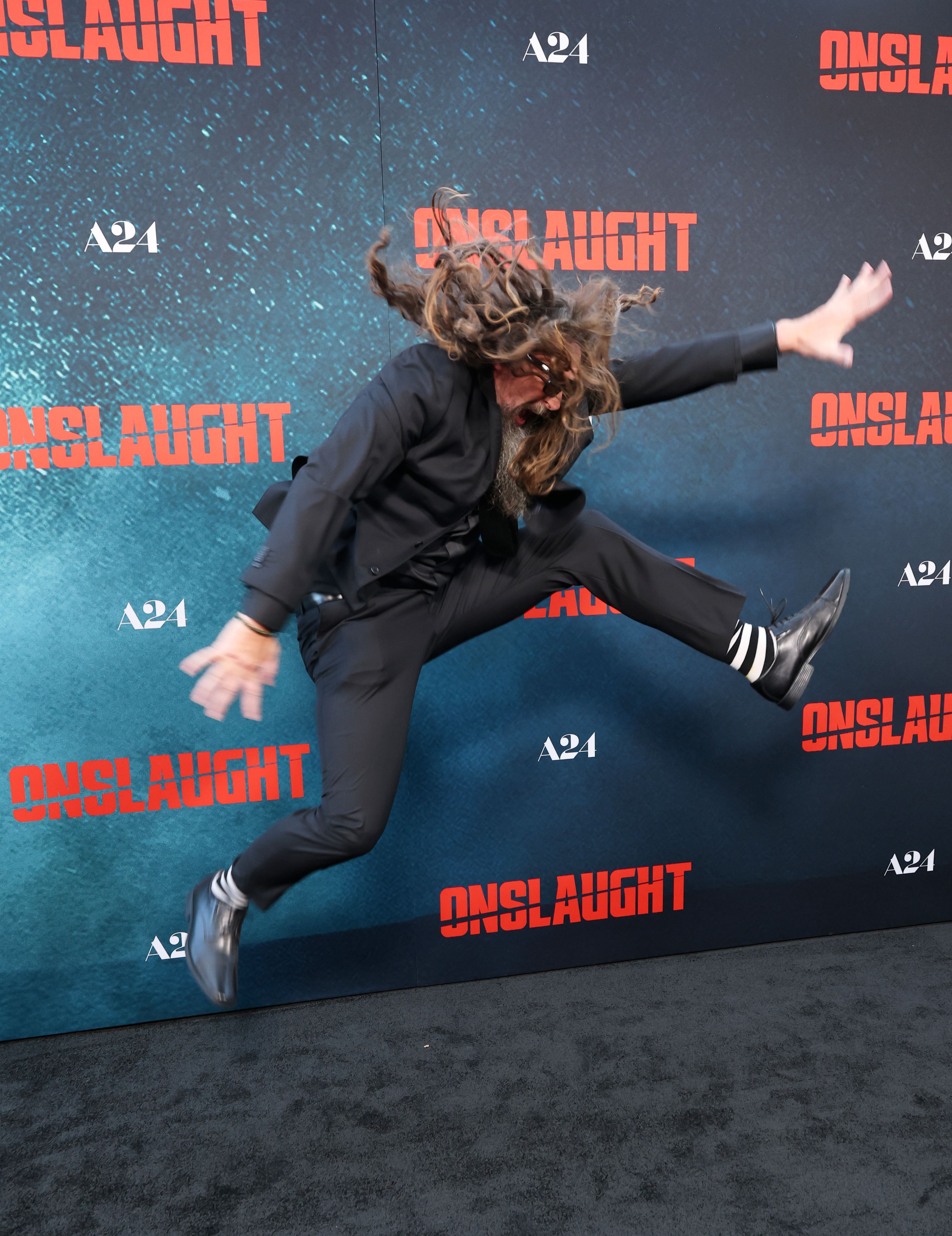 Adam Wingard jumping in the air, arms and legs spread wide, long hair flying, in front of an A24 Onslaught premiere backdrop.